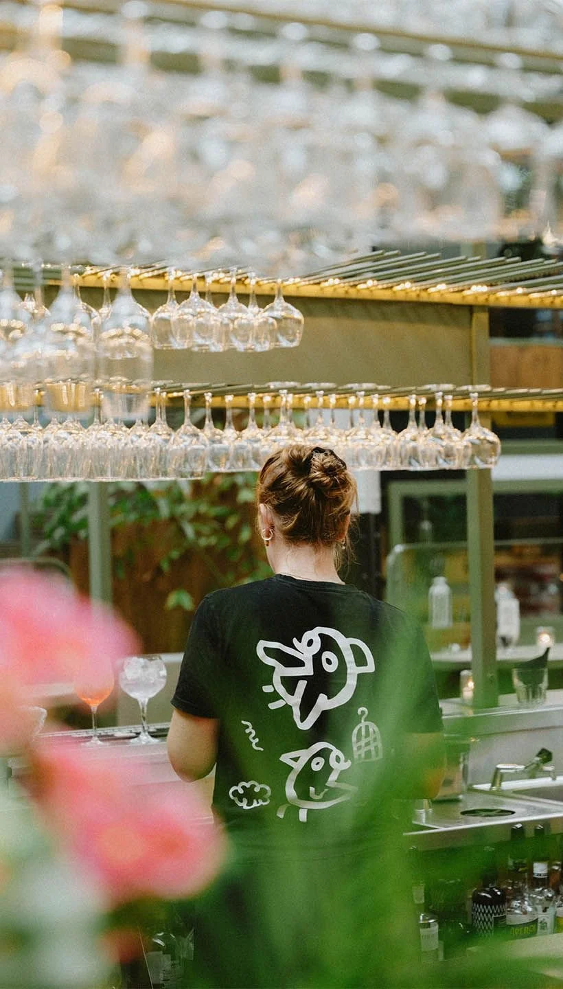 A woman with red hair tied up is seen from behind, working at a bar or restaurant with hanging wine glasses above her.