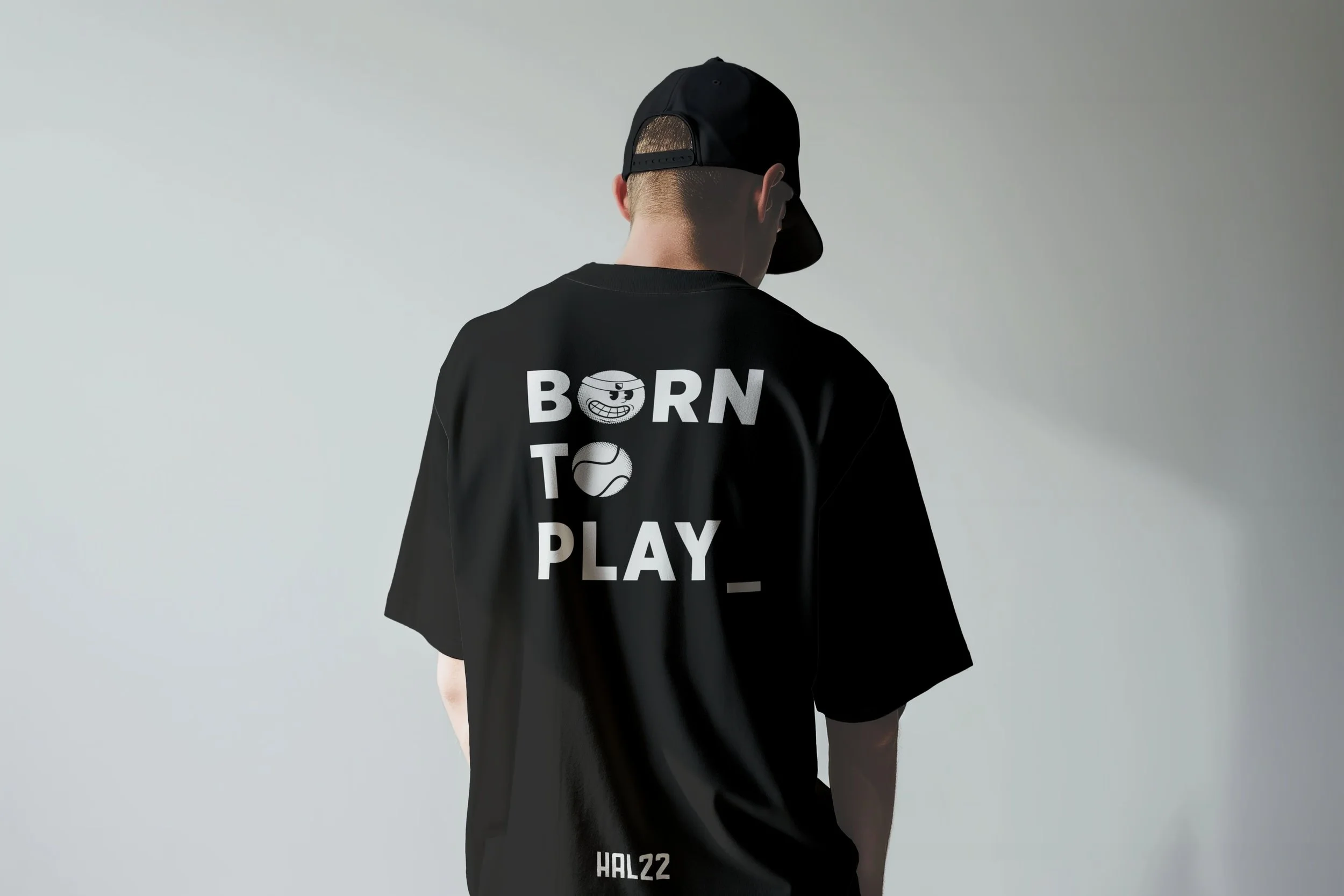 Back of a person wearing a black T-shirt with the text 'BORN TO PLAY' and a graphic of a smiling ball, along with a black cap, standing against a plain gray wall.