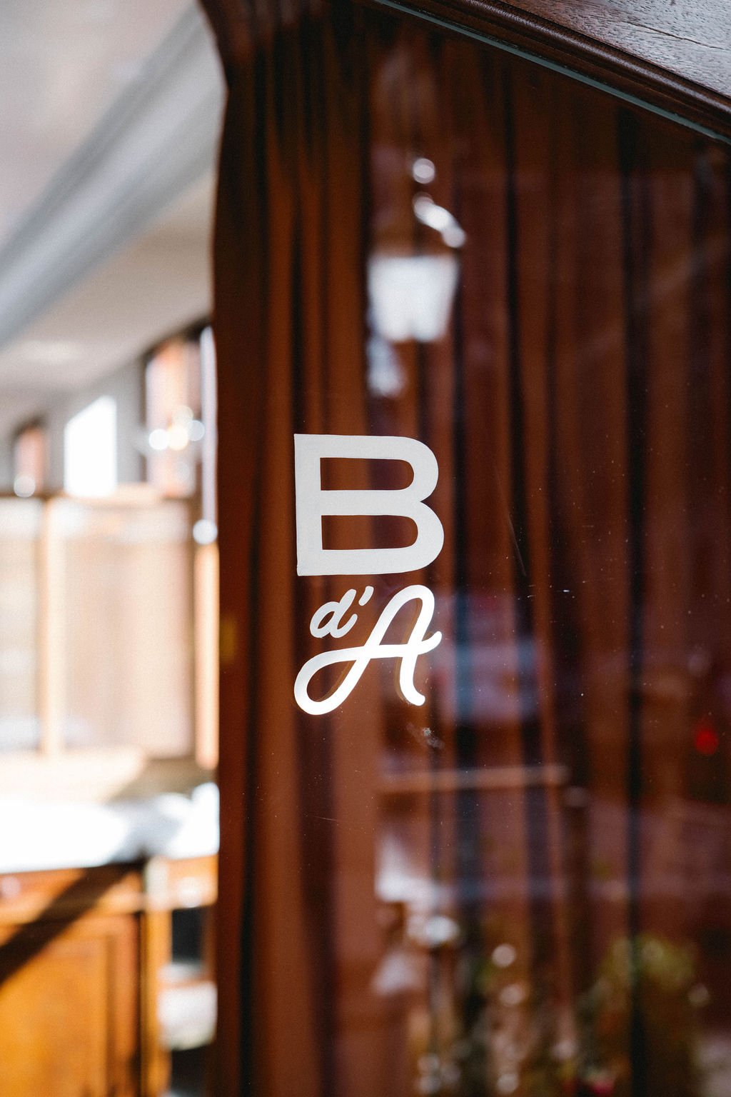Close-up of a glass door with a white sign that reads "B d'A" in a stylish font, with blurred interior background and warm lighting.