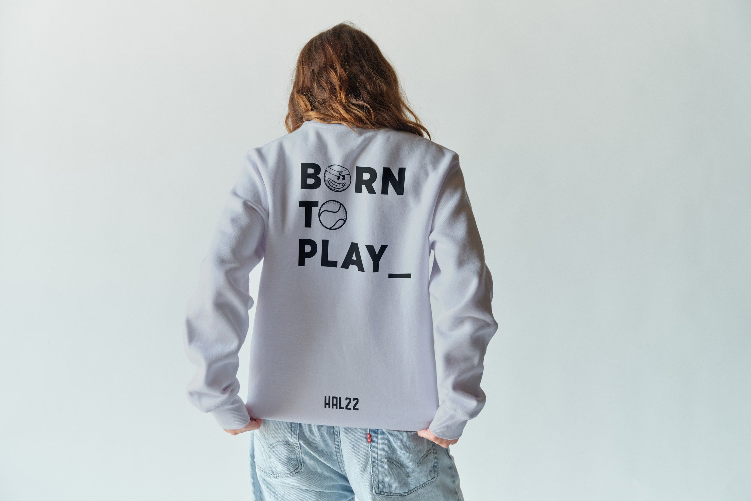 Back view of a person with brown wavy hair wearing a white sweatshirt with 'BORN TO PLAY_' printed on the back and light blue jeans.