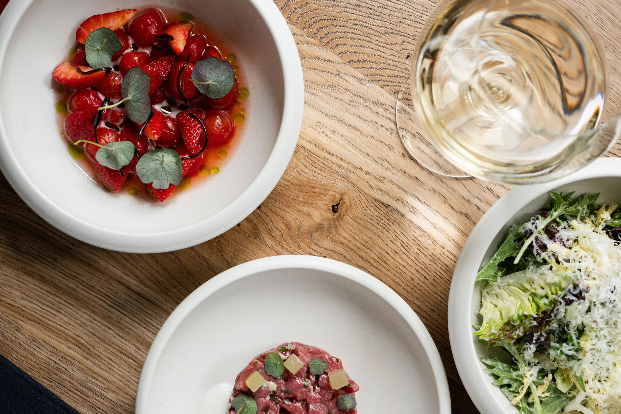 A top view of a wooden table with a glass of white wine, a bowl of mixed salad with cheese, a bowl of fresh strawberries and cherries, and a plate of beef tartare topped with small cheese cubes and greens.