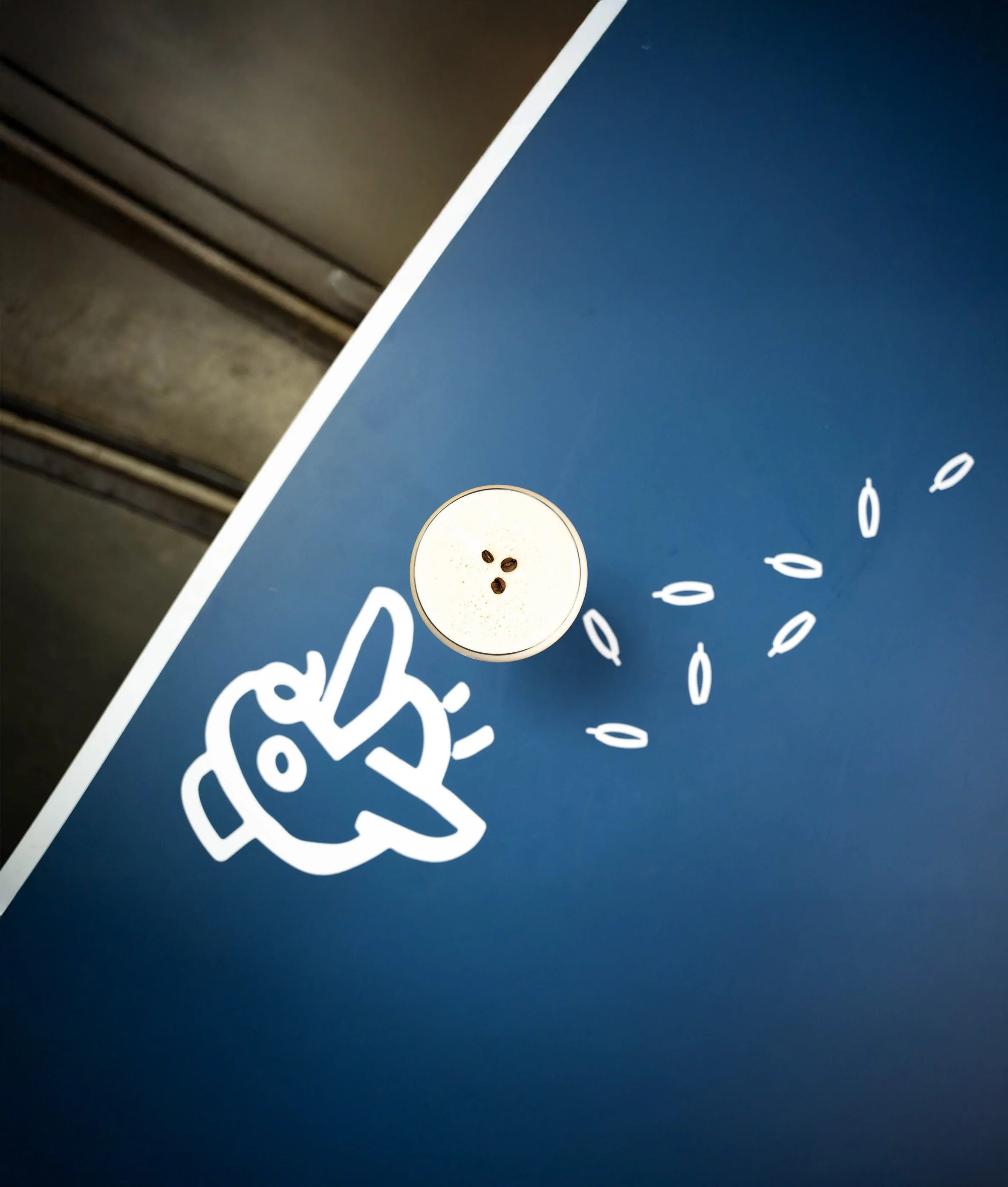 A top-down view of a blue table with a drink topped with foam and coffee beans, a white doodle of a hand making a peace sign, and small shapes resembling seeds or grains.