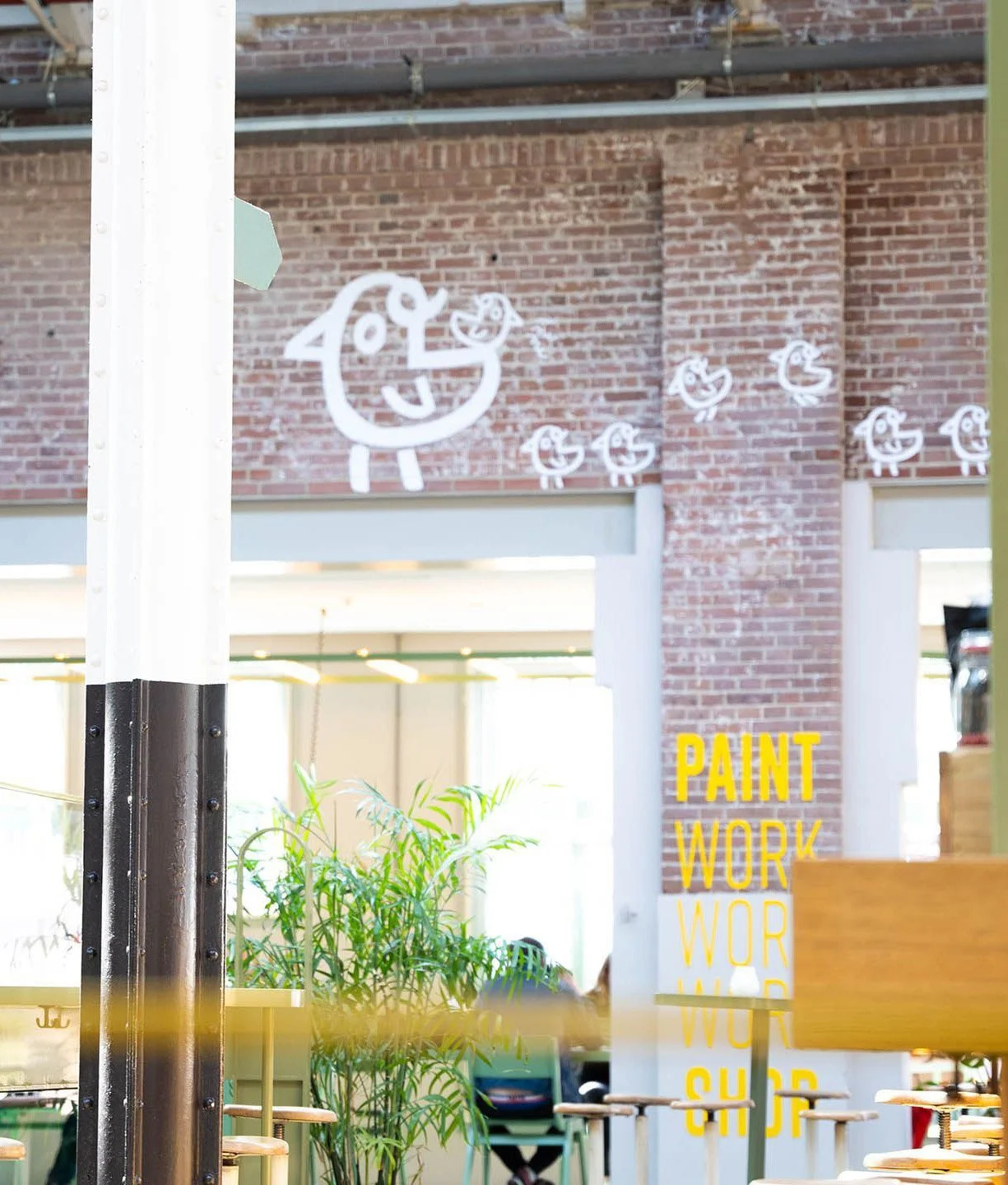 Decorative sign with a smiling cartoon chicken and smaller chickens on a brick wall in a restaurant or cafe with plants and yellow text that reads 'PAINT WORK'.