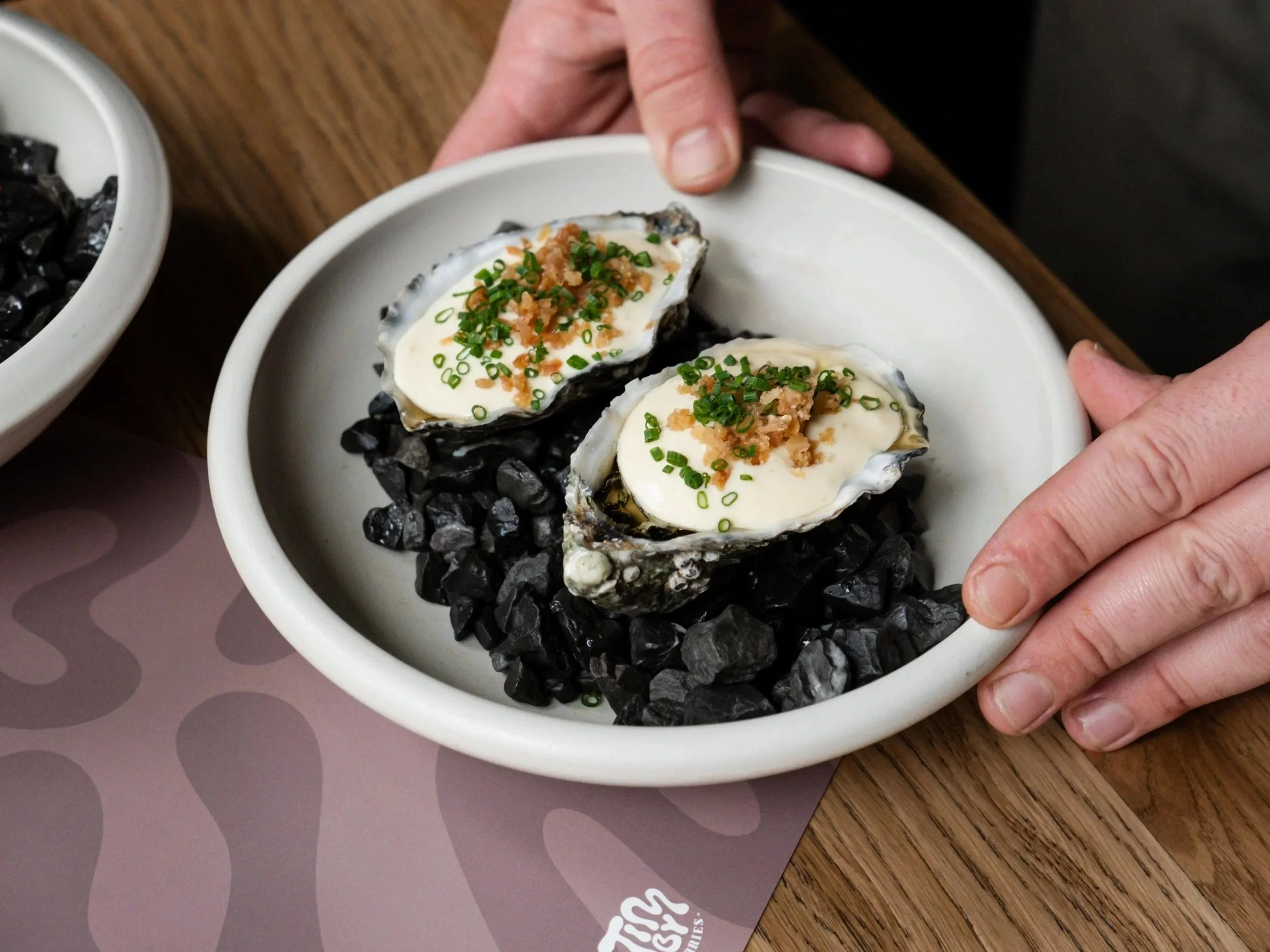 Oysters topped with cream, chopped green onions, and crunchy bits, served on black rocks in a white bowl.