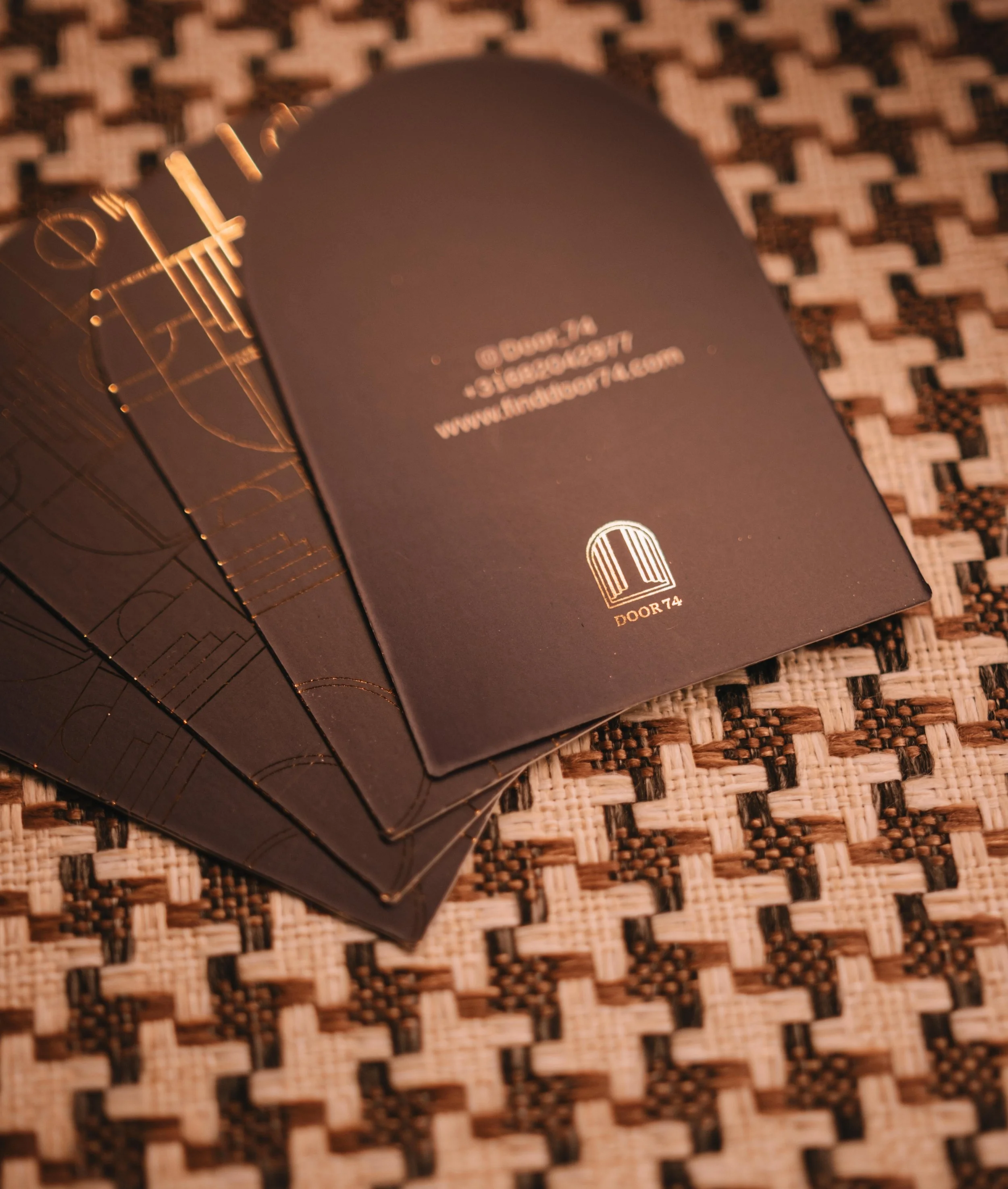 A set of black business cards with gold foil accents, stacked on a woven surface. The top card displays a logo and contact information for DOOR74.