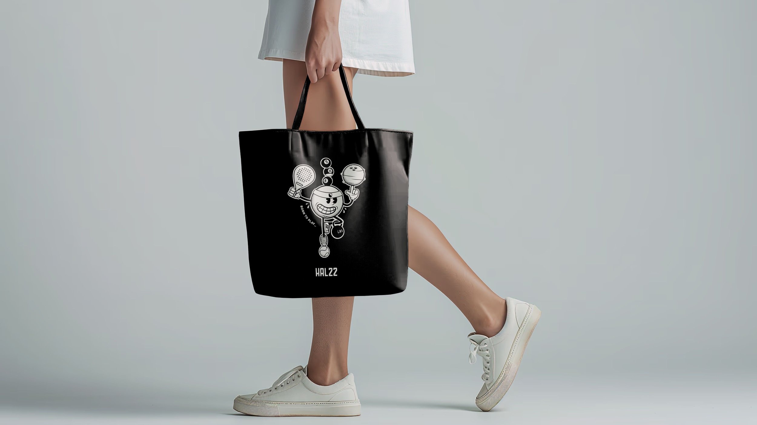 Person wearing white sneakers and a light dress holding a black tote bag with a cartoon tennis-playing character and the text "HAL22" on it, standing against a plain light gray background.