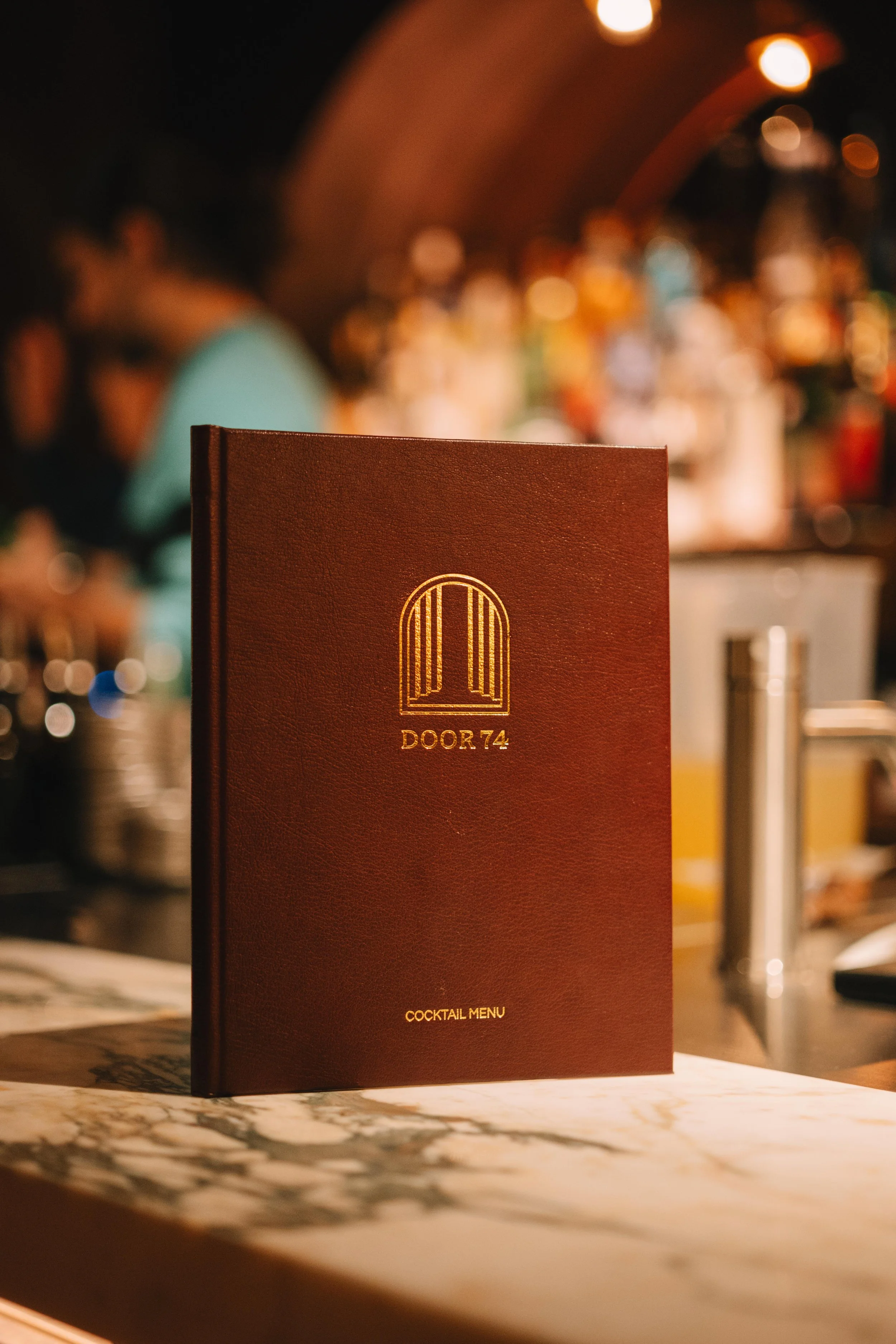 A cocktail menu with a dark red cover featuring a gold arch logo and the words 'DOOR 74' and 'COCKTAIL MENU' on a bar counter, with a blurred background of bar shelves and patrons.