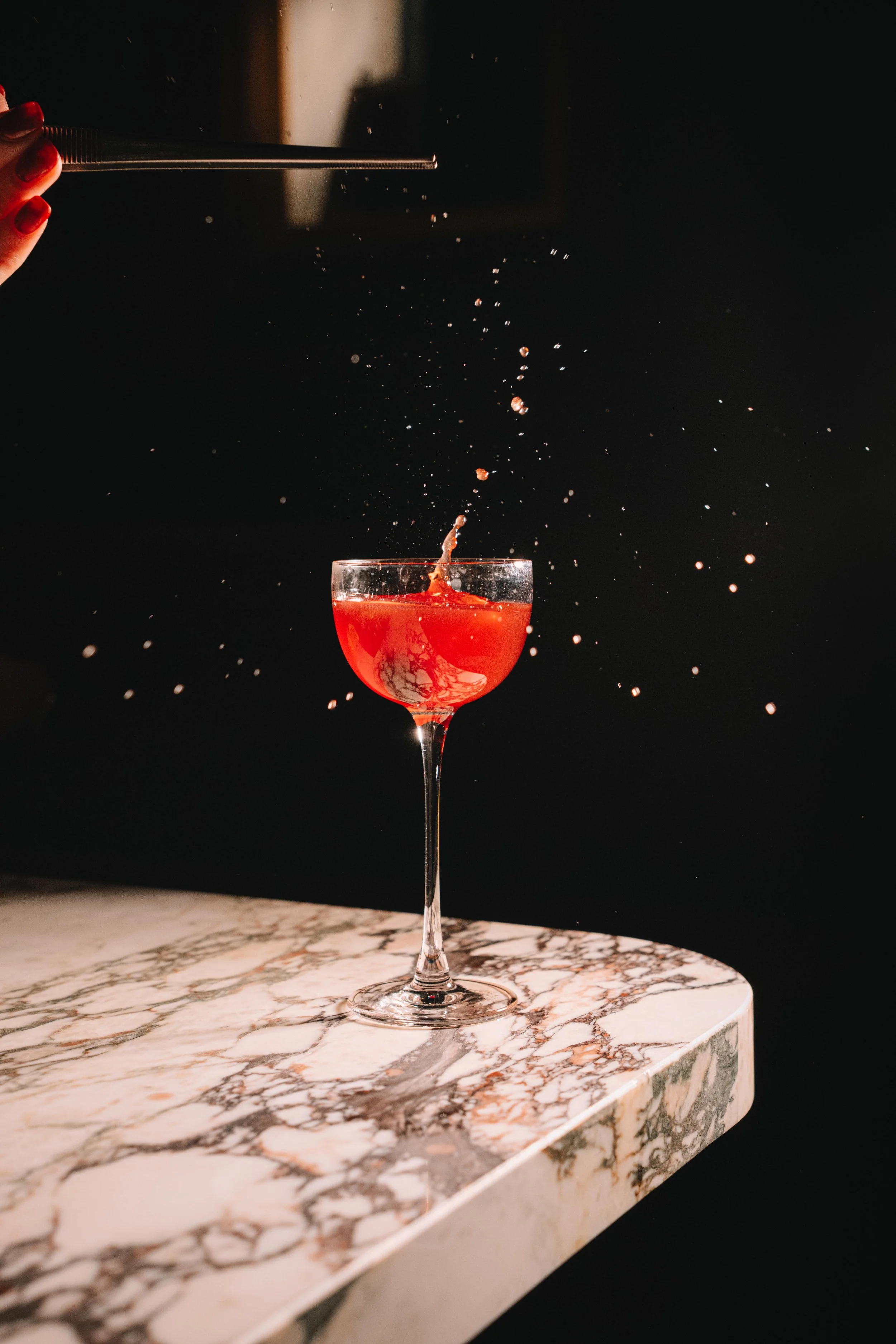 A hand with red manicured nails is pouring a drink into a cocktail glass with a long stem, placed on a marble table. The drink is red, and the liquid is splashing as it is poured into the glass against a dark background.