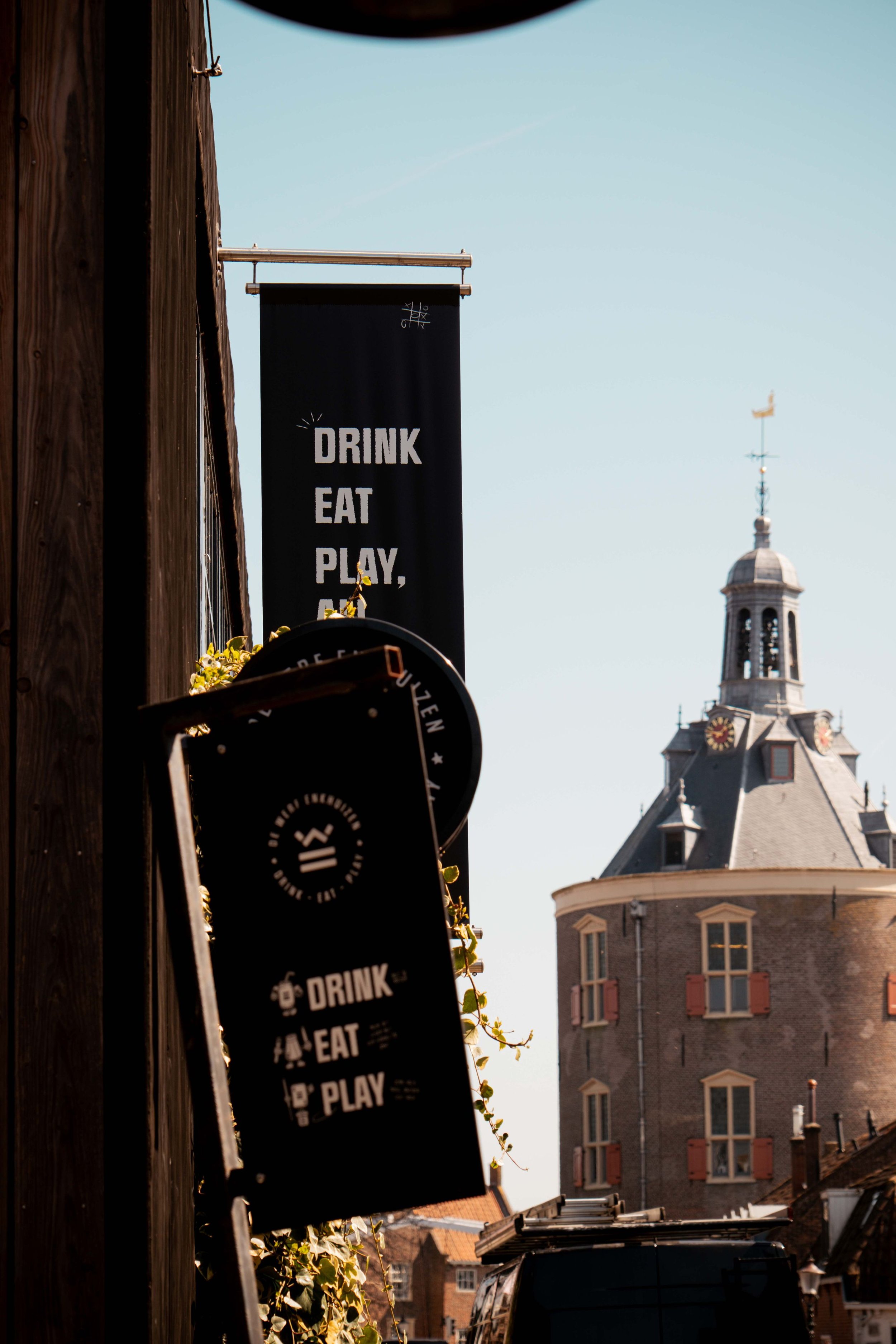 Street view with hanging signs that read "Drink, Eat, Play" and a historic building with a clock tower in the background.