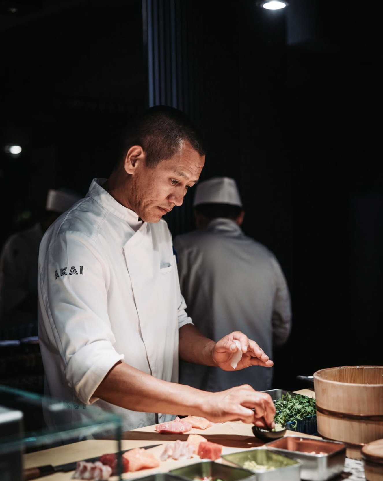 A chef preparing sushi in a dimly lit kitchen, with other chefs in the background.