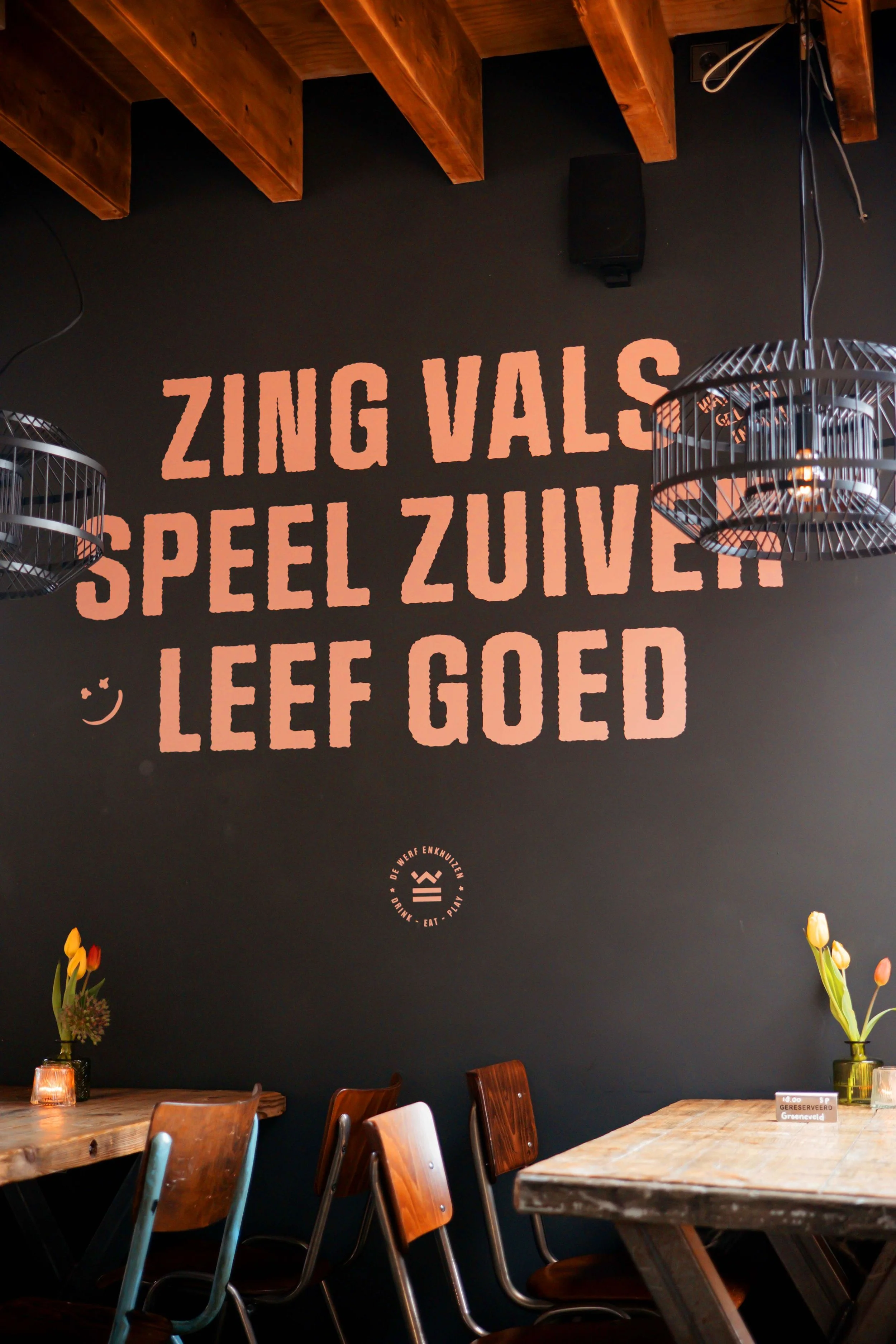 Interior of a restaurant with a dark wall and wooden ceiling beams, decorated with hanging light fixtures, flowers in vases on wooden tables, and a large mural with Dutch words that say 'Zing Vals, Speel Zeker, Leef Goed'.