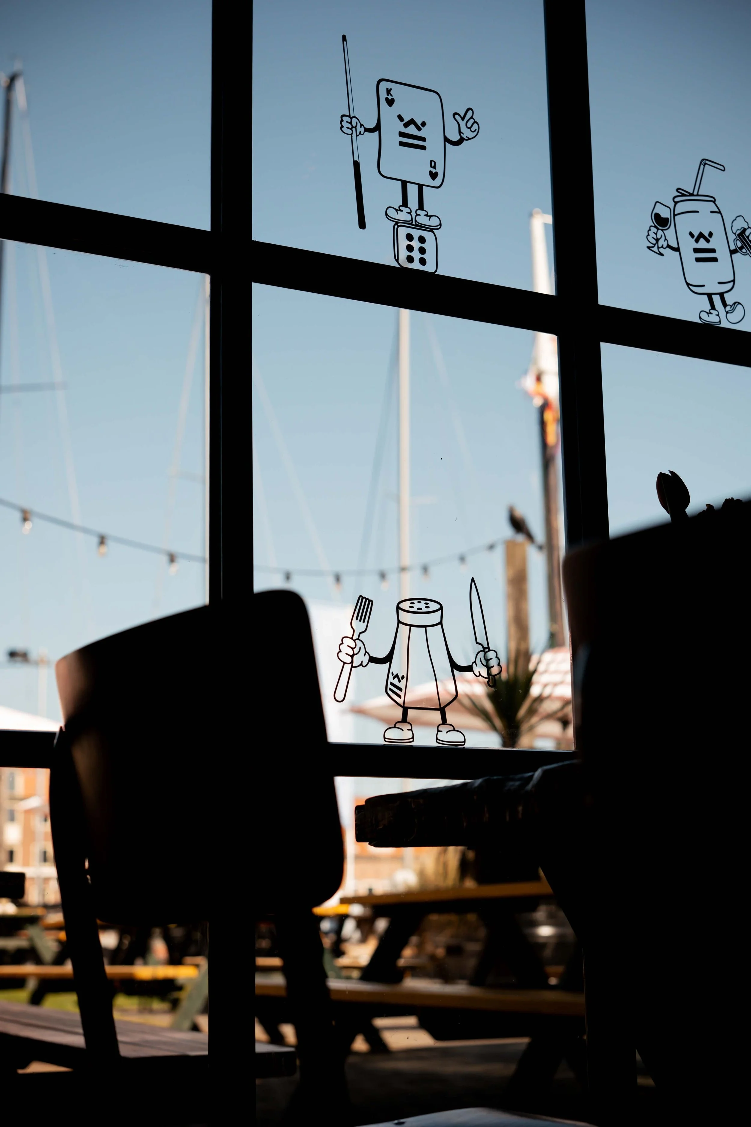 Decorative stickers of cartoon restaurant utensils and drink characters on a window at a boarded-up outdoor seating area, with reflections of sailboats and a crane outside.
