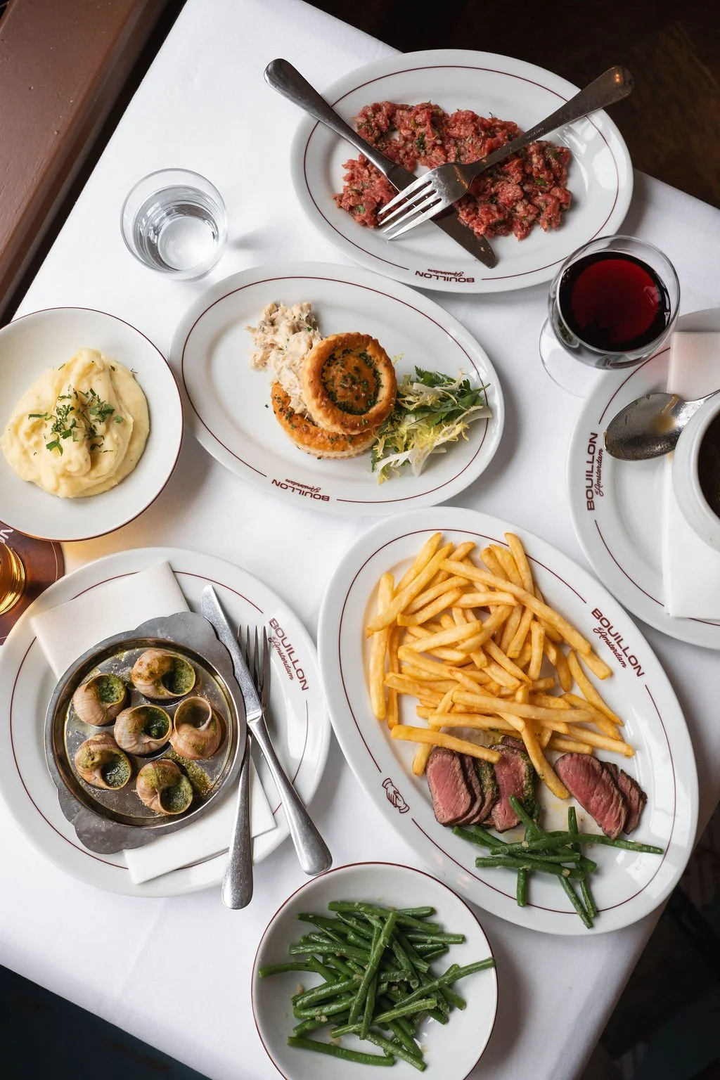 A table set with various dishes including mashed potatoes, escargot, sliced steak with fries and green beans, a breaded patty with sauce, a bowl of shellfish, steak tartare, a glass of red wine, a glass of water, and a cup of coffee.