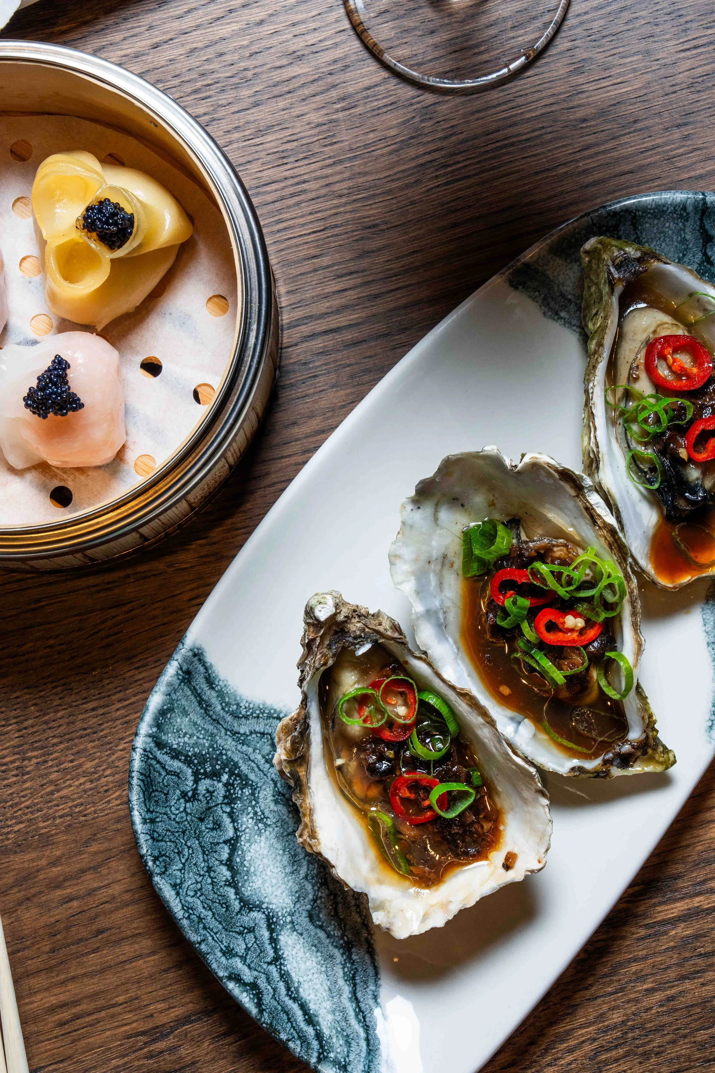Three oysters topped with sauce, sliced red and green chilies, and green onions on a white and blue plate, and two steamed dumplings with black caviar in a bamboo steamer.