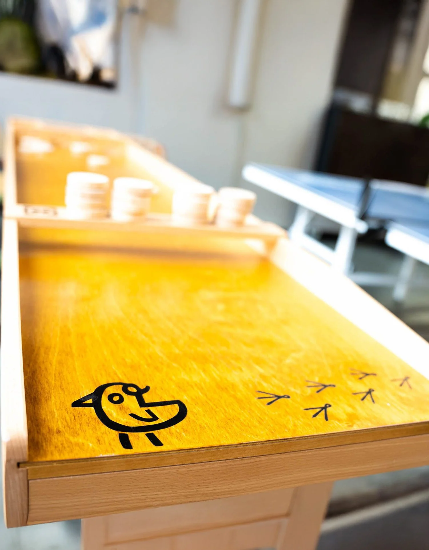 Wooden shuffleboard game with a cartoon chick sticker and bird footprints, blurred background with white cups.