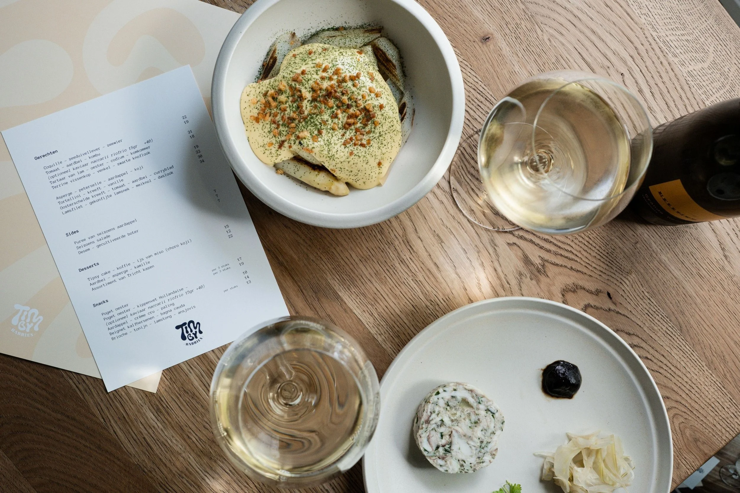 Table setting with two glasses of white wine, a menu, a bowl with fish covered in sauce, and a plate with a piece of cheese, a small black item, and some sliced onions, on a wooden table.