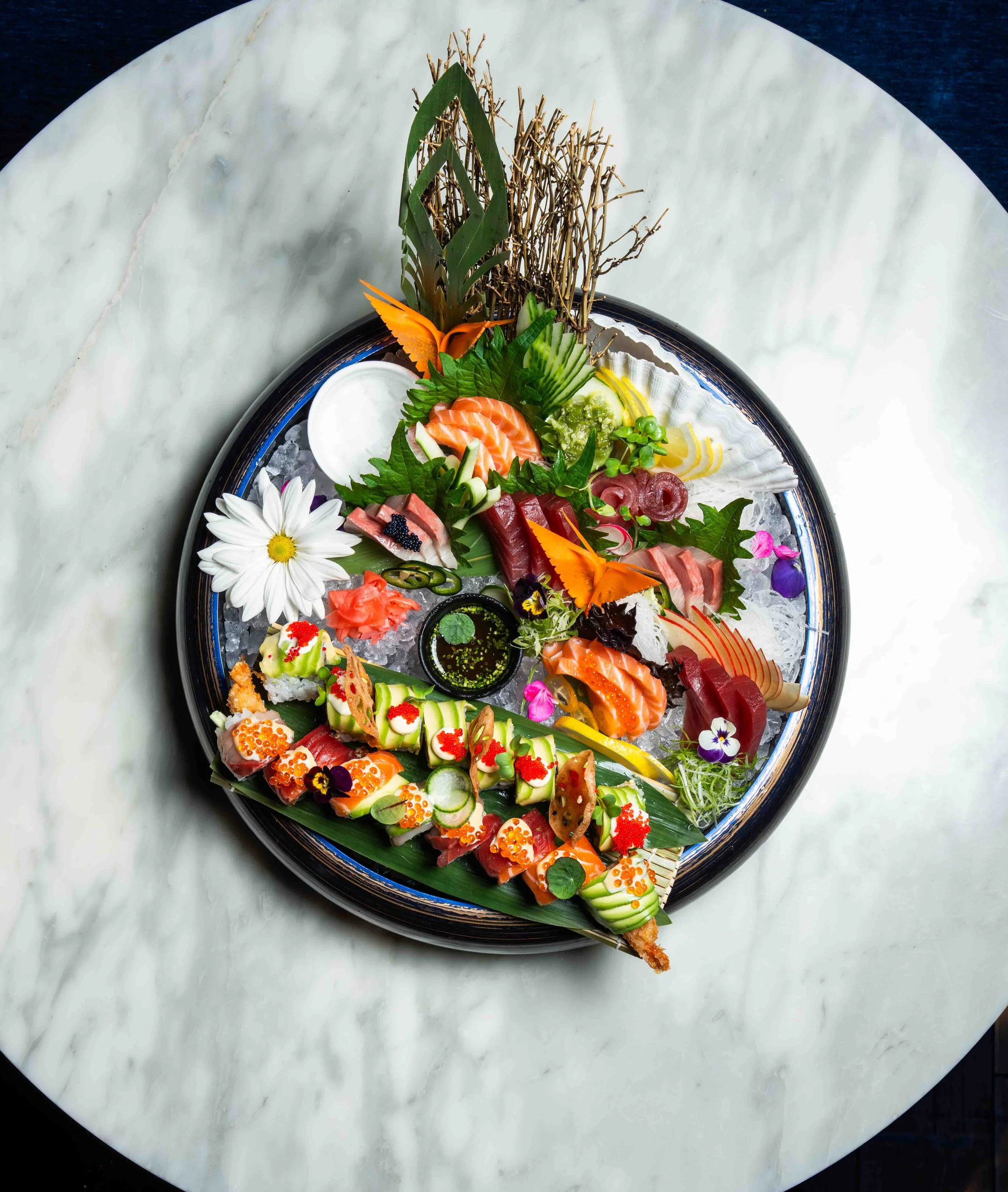 A round platter of assorted sashimi and sushi on ice, garnished with edible flowers, cucumber slices, and decorative leaves, with a small container of soy sauce in the center.