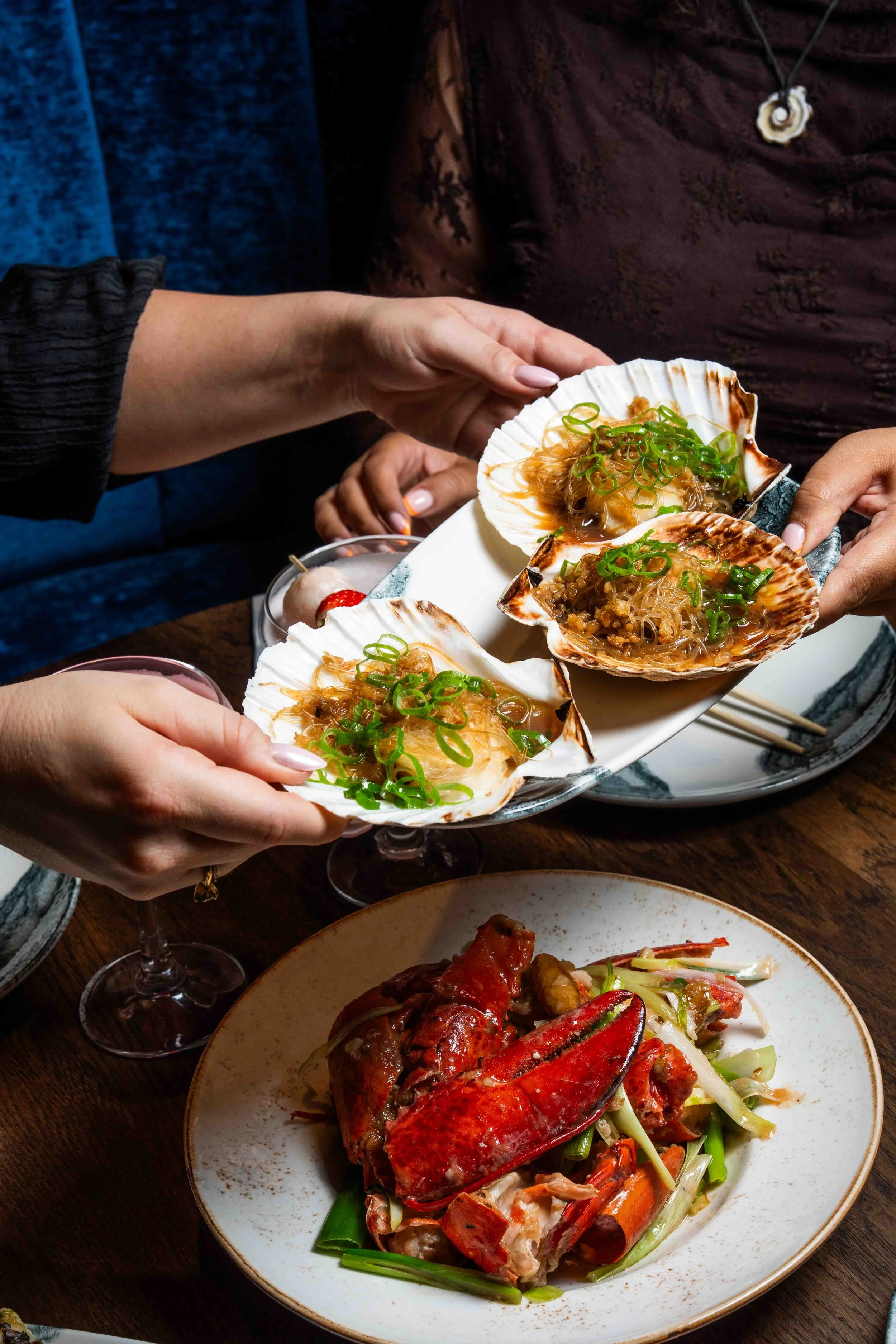 People serving and sharing scallops topped with green onions at a restaurant table, with a lobster and vegetable dish in the foreground.