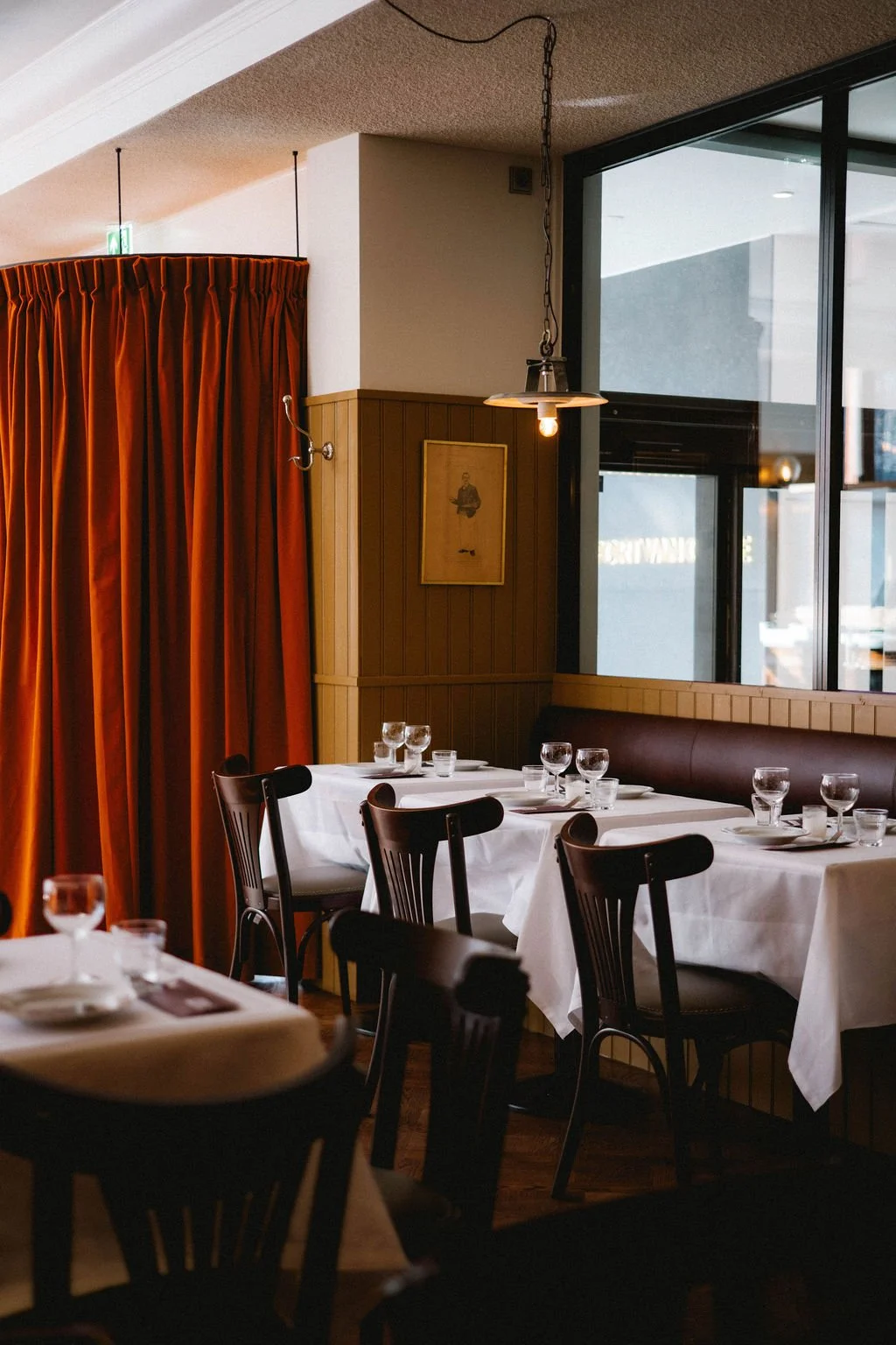 Empty restaurant dining area with white tablecloths, glassware, dark wooden chairs, large window, and orange curtains.