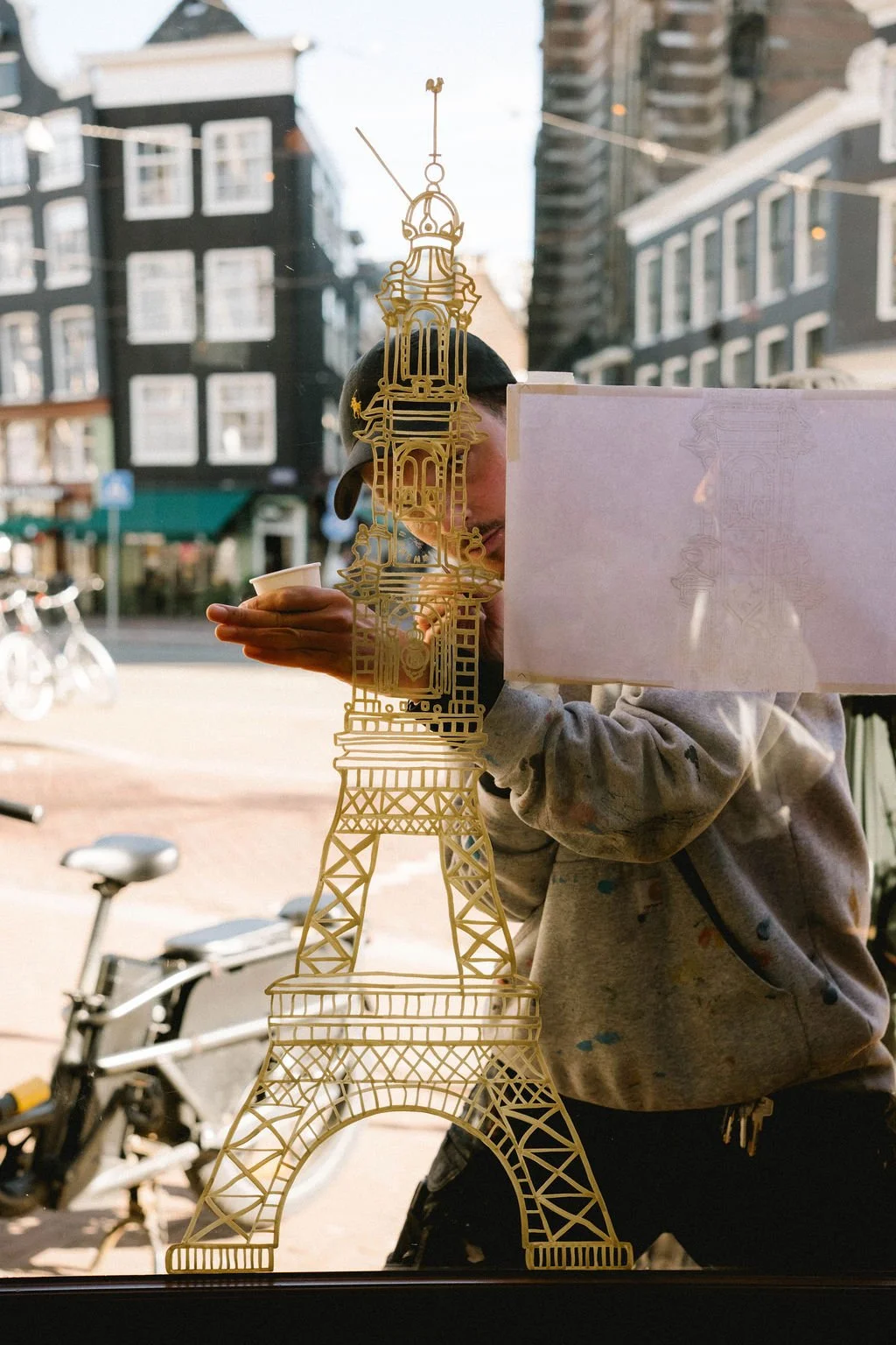 Person holding a cup of coffee, seen through a glass window decorated with a gold outline of the Eiffel Tower, with buildings and bicycles outside.