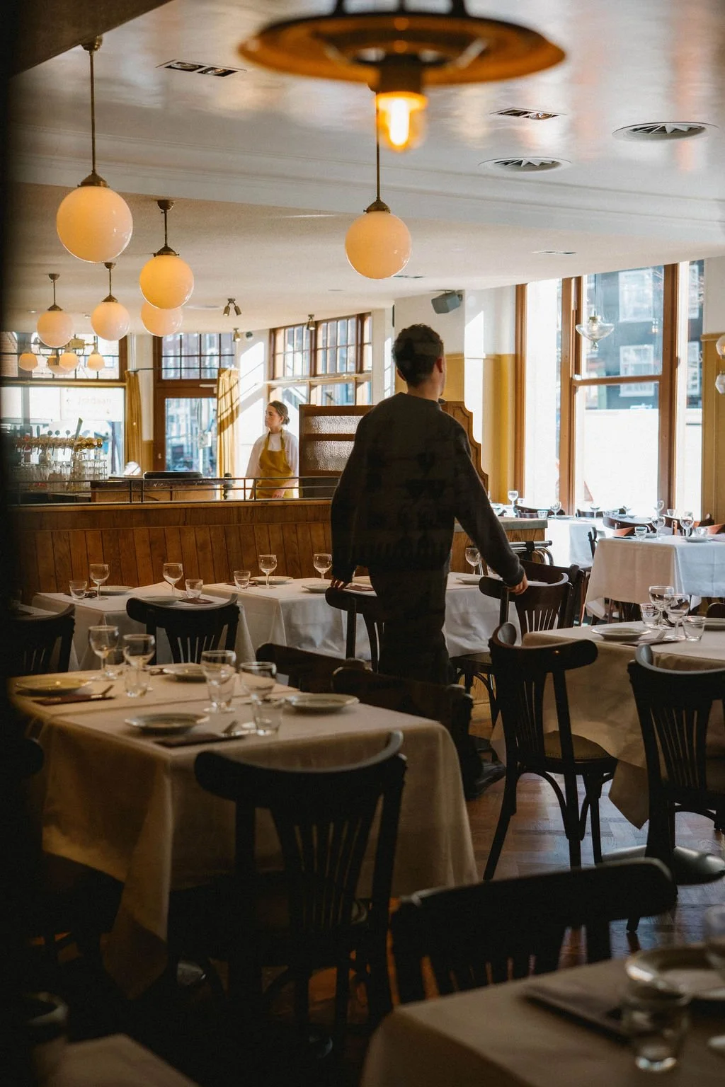 A man in a dark shirt pushes a trolley through a well-lit restaurant with white tablecloths, glassware, and black chairs, with a woman in a yellow apron standing near the entrance.