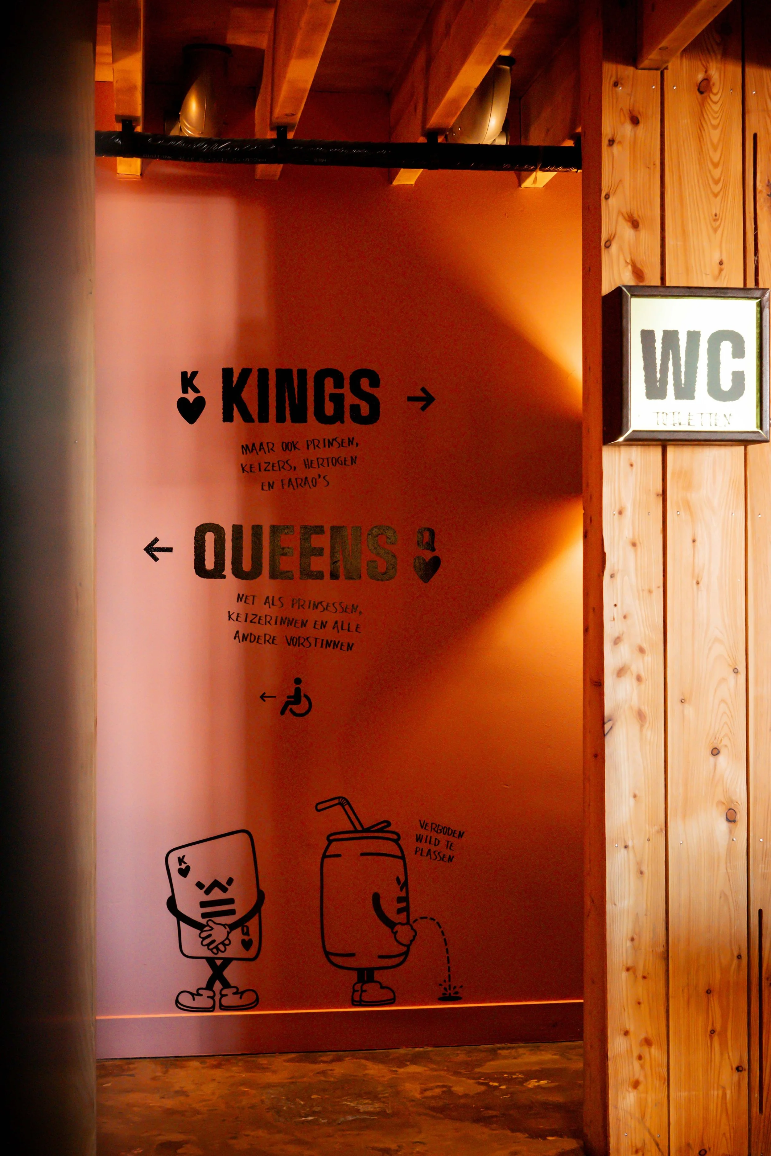 A sign on a pink wall with black text and illustrations. It indicates directions to the 'Kings' to the right and 'Queens' to the left. The sign includes small handwritten notes in Dutch and drawings of animated characters, one holding a card and the other watering a plant, with a 'WC' sign nearby.