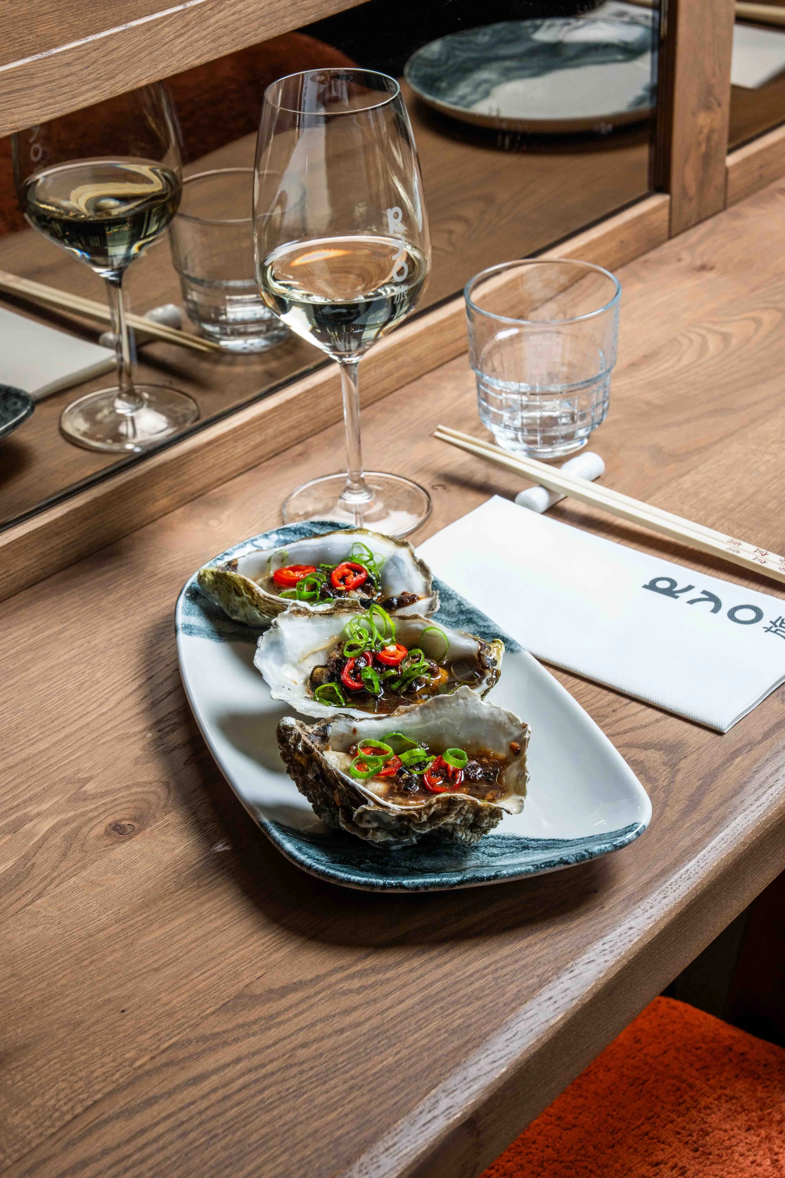 A plate of three oysters garnished with sliced red and green chili peppers and chopped green onions, on a white and blue ceramic dish on a wooden table. Two glasses of white wine and a glass of water are in the background, along with a mirror reflecting the scene.