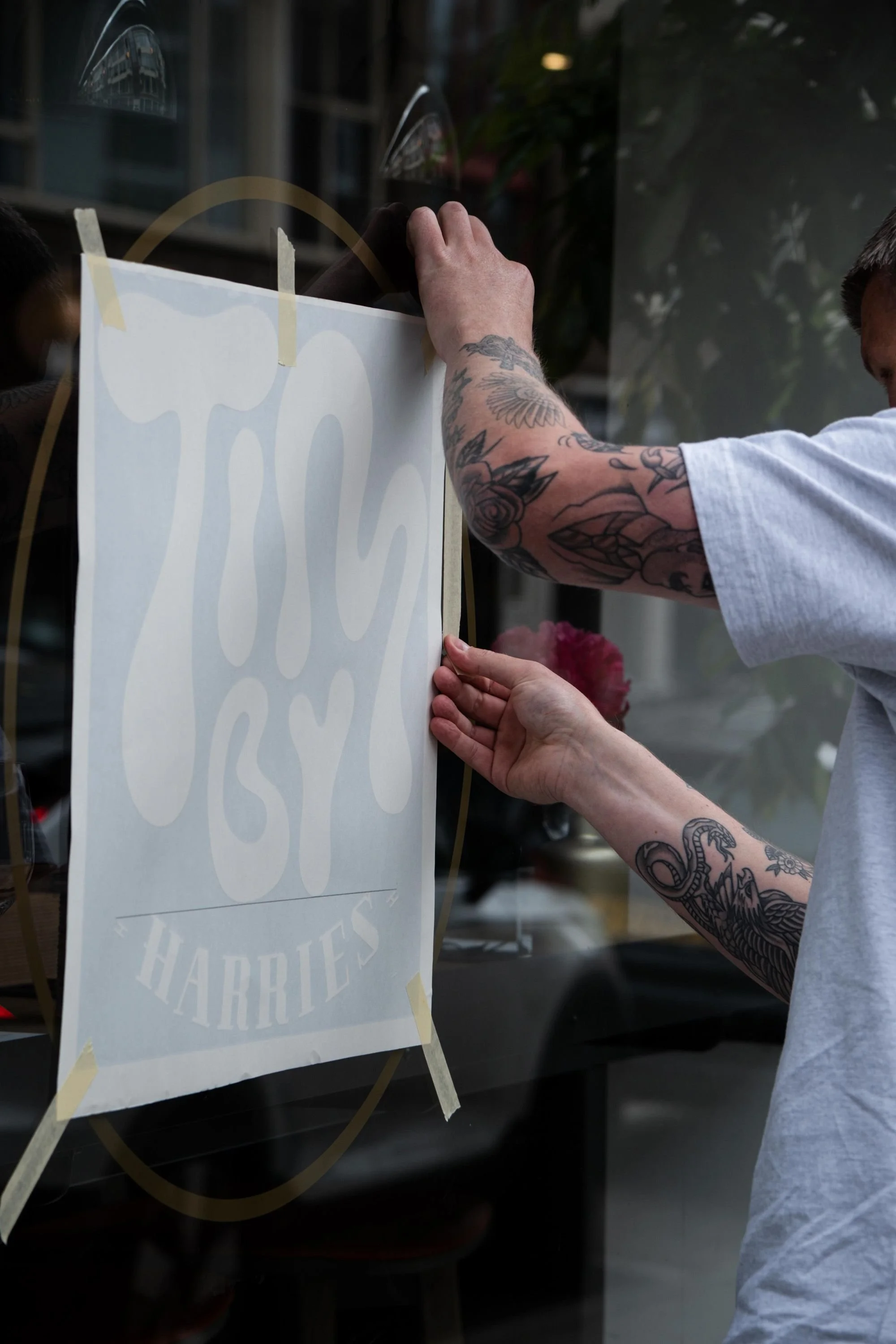 Person applying a sign to a window with masking tape, tattooed arms, and a white t-shirt.