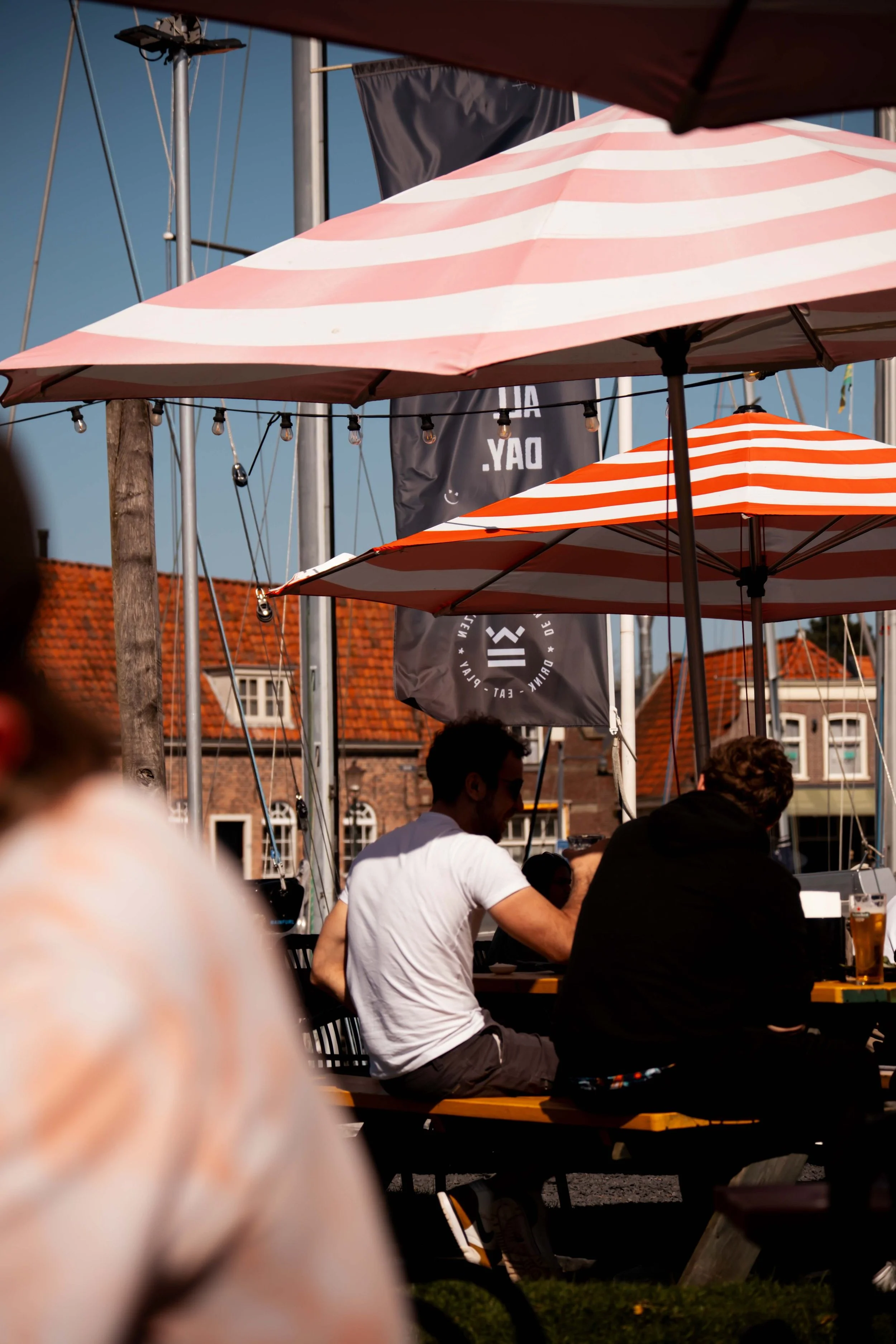 People sitting at outdoor tables under pink and orange striped umbrellas at a sunny patio during daytime, with buildings and flags in the background.
