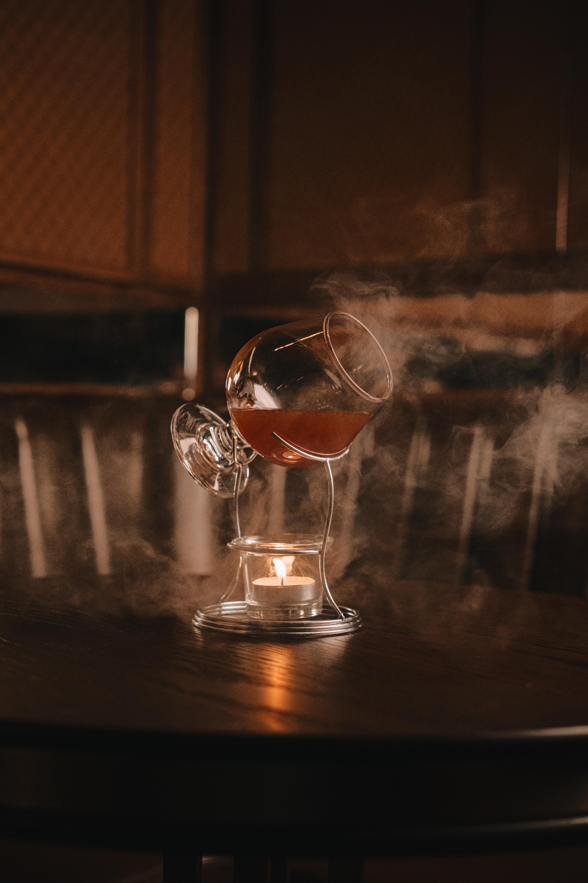 A glass of dark liquid in a tilted glass cup with steam and a lit candle underneath, set on a dark wooden table.