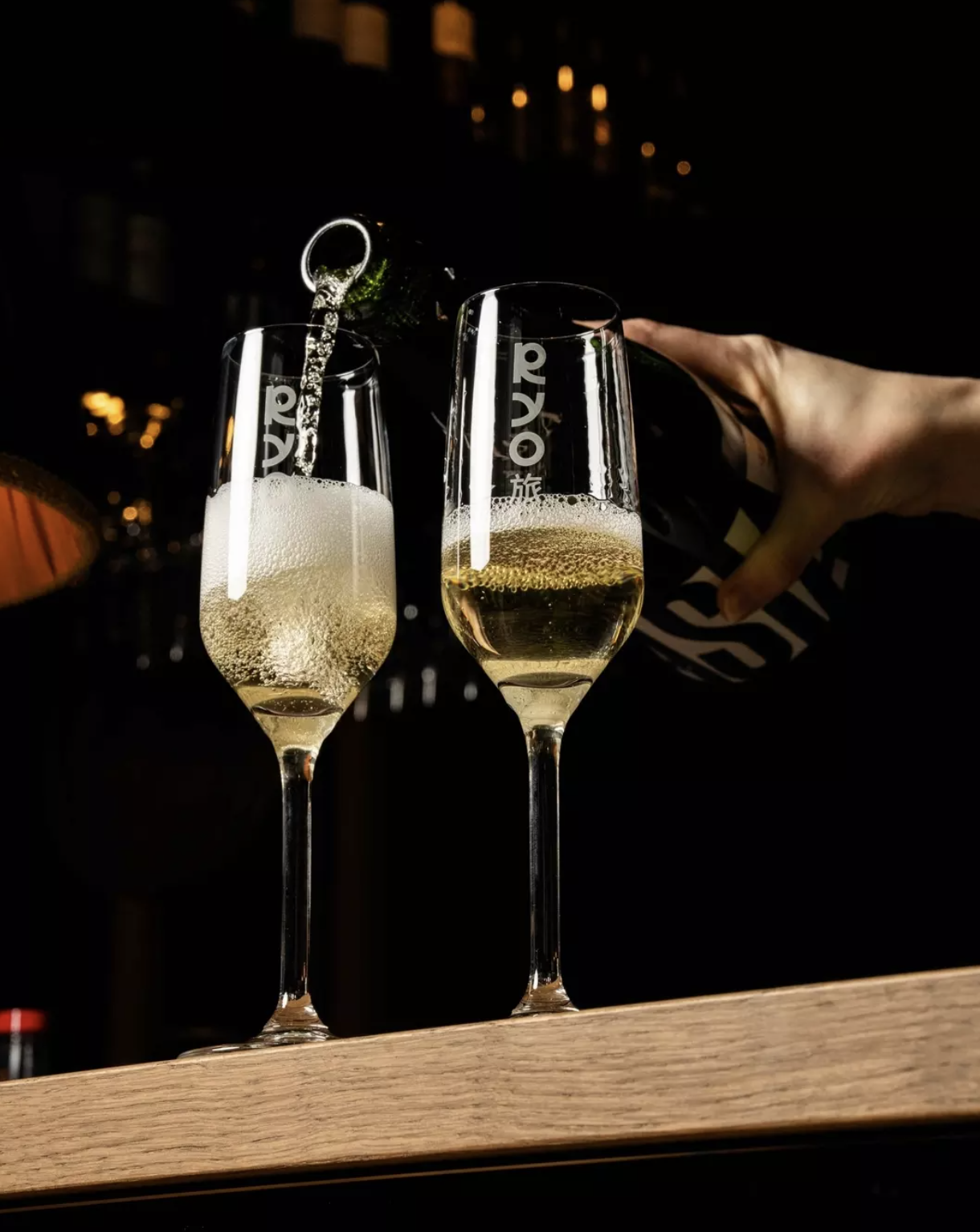 A person pouring champagne into two glasses on a wooden surface in a dark setting.