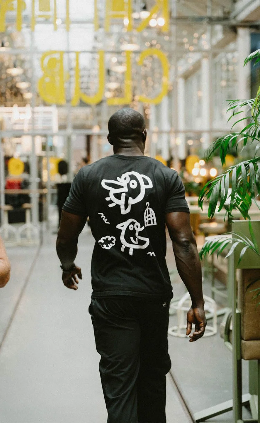 Back view of a man walking through a modern, brightly lit indoor space, possibly a cafe or co-working area, with green plants and glass walls.