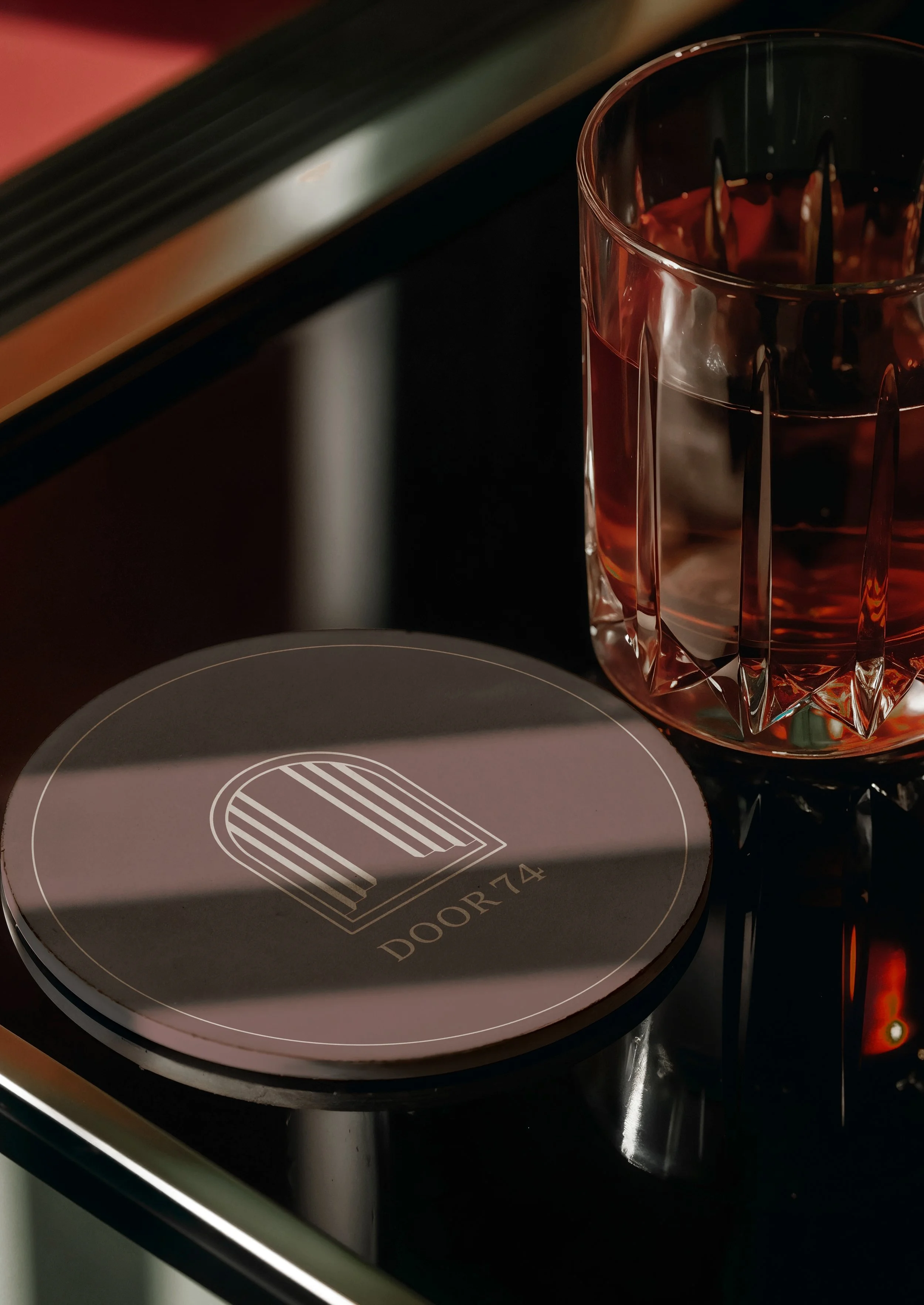 A round, modern coaster with the logo "DOOR74" placed next to a cut-glass tumbler filled with a reddish beverage, on a black surface with a shadow cast across the items.