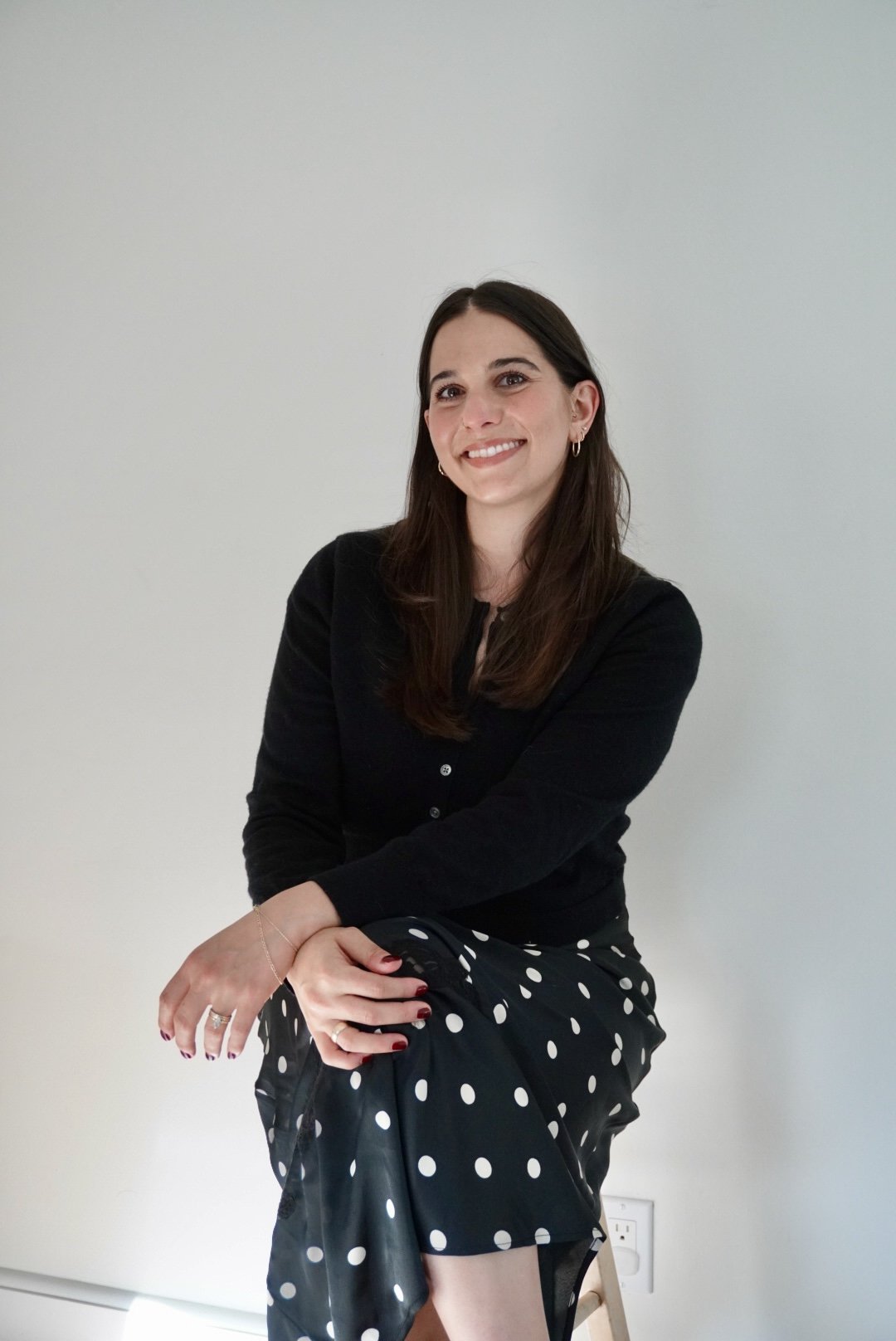 A woman with long brown hair wearing a black cardigan and a polka dot skirt, smiling and sitting on a stool against a light-colored wall.