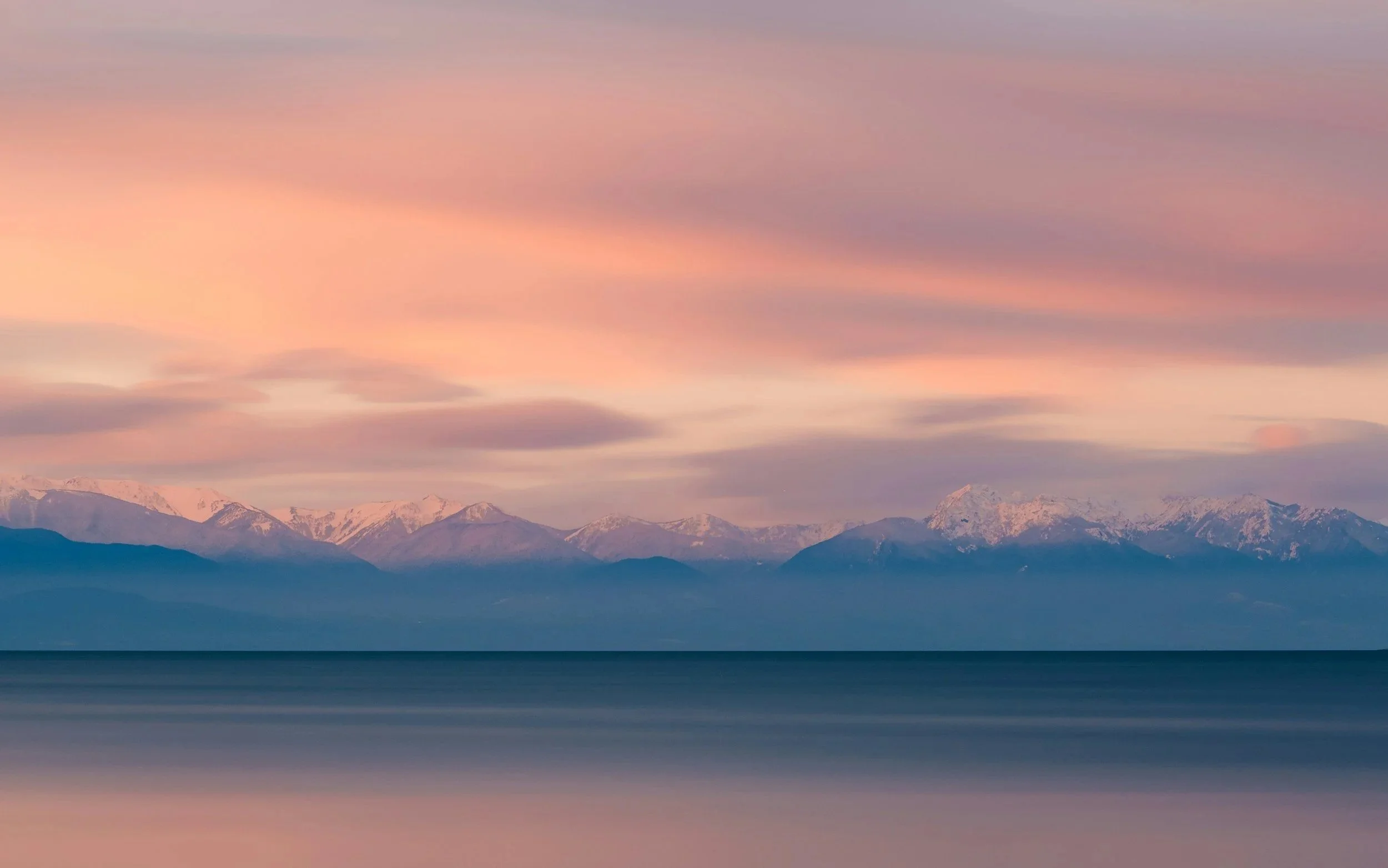 A serene landscape featuring snow-capped mountains in the background under a pink and purple sky with wispy clouds, reflected on a calm body of water in the foreground.