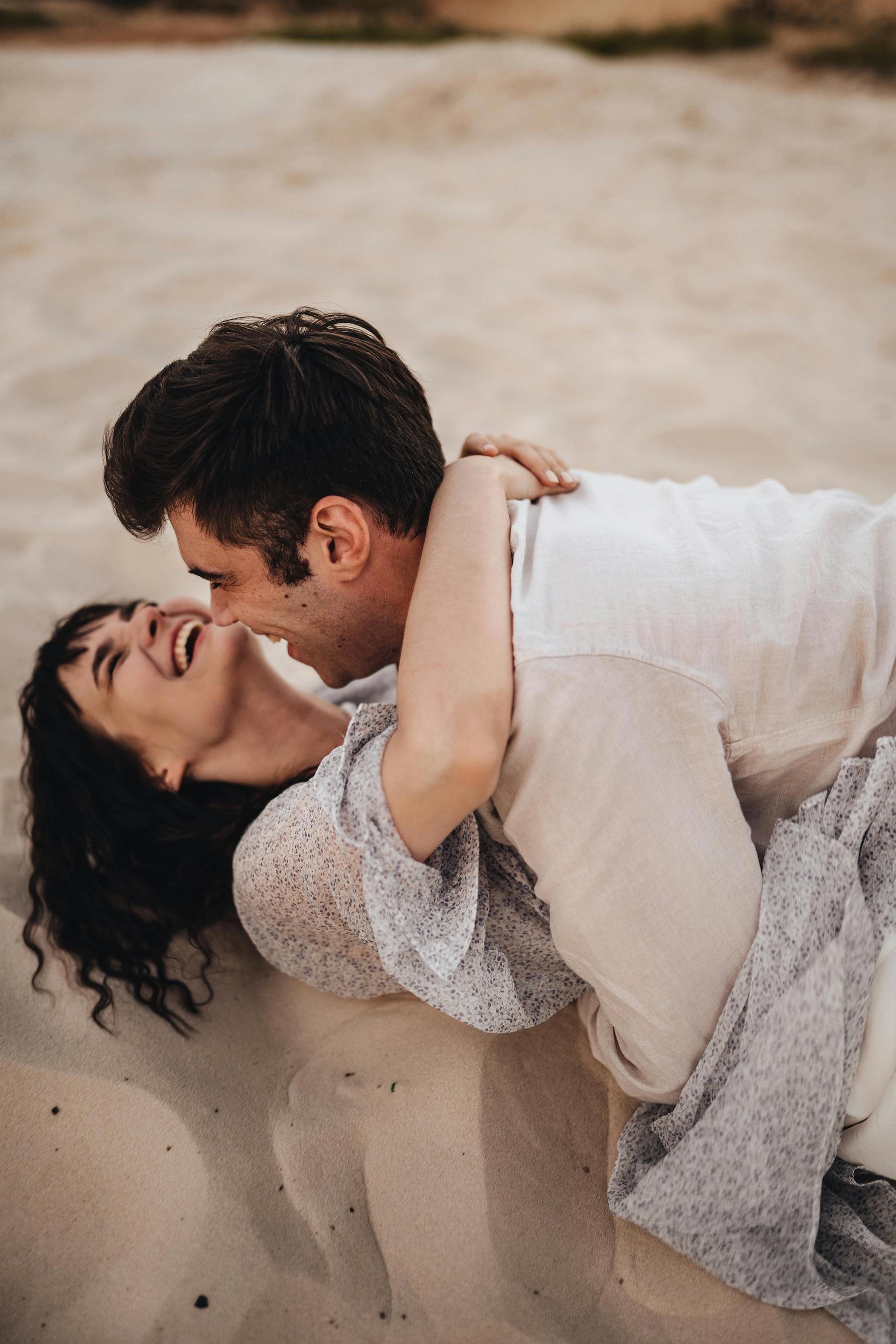 A man and a woman are lying on a sandy beach, laughing and embracing each other.