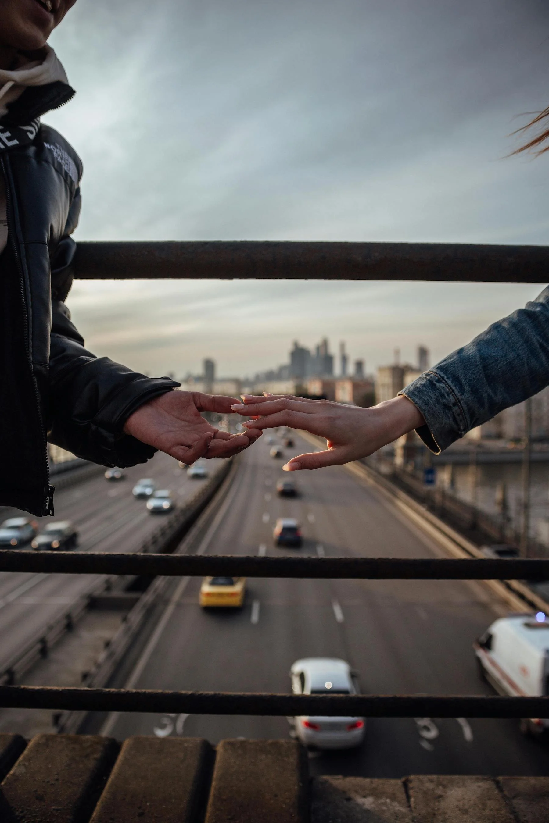 Close-up of a man and a woman reaching out their hands towards each other on a bridge with a cityscape and highway below in the background.
