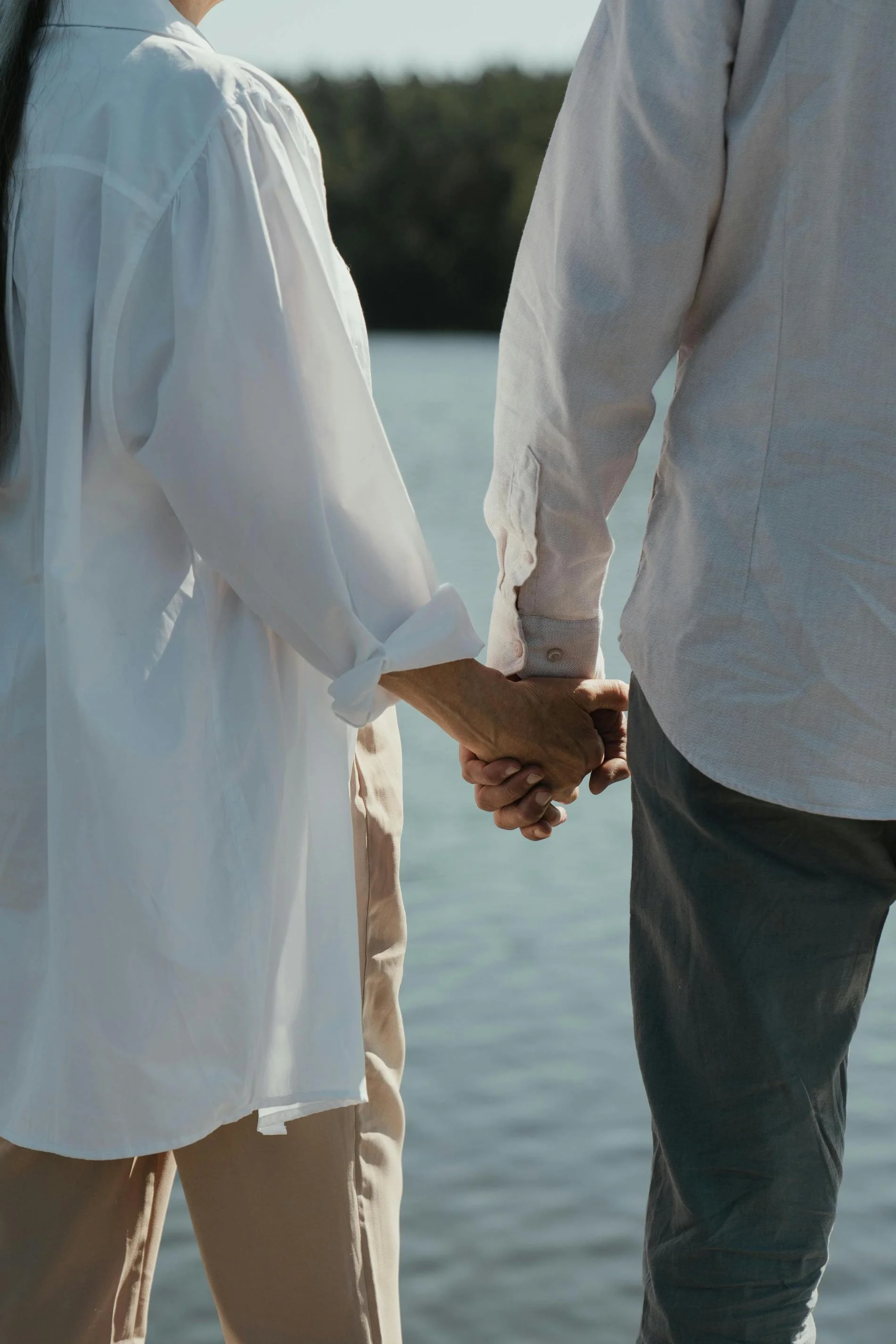 Close-up of a couple holding hands near a body of water, with trees in the background.