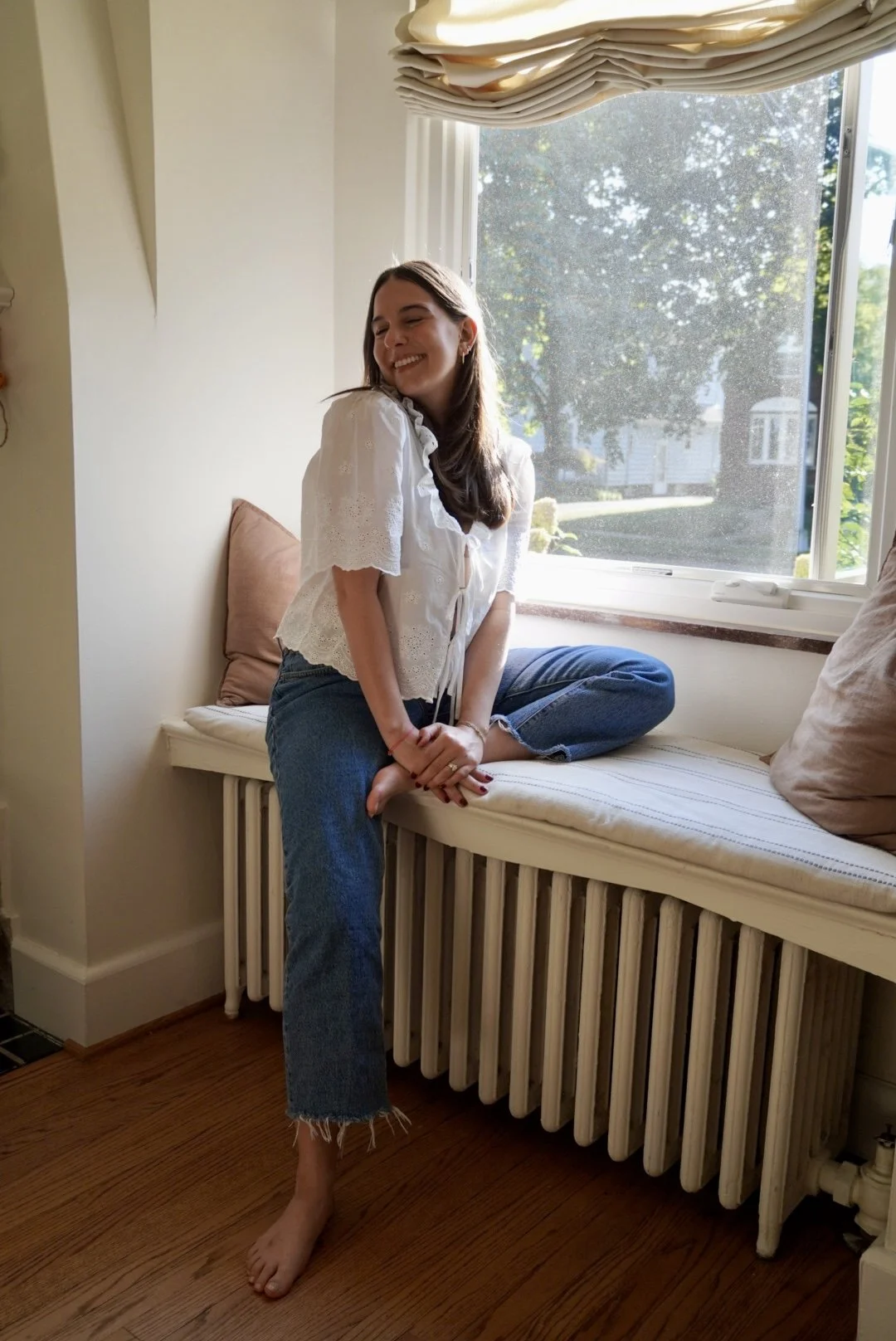 A woman sitting on a window seat with her legs crossed, smiling with her eyes closed. She's wearing a white blouse with lace sleeves and blue jeans with frayed hems. Sunlight streams through the window behind her, illuminating the room.