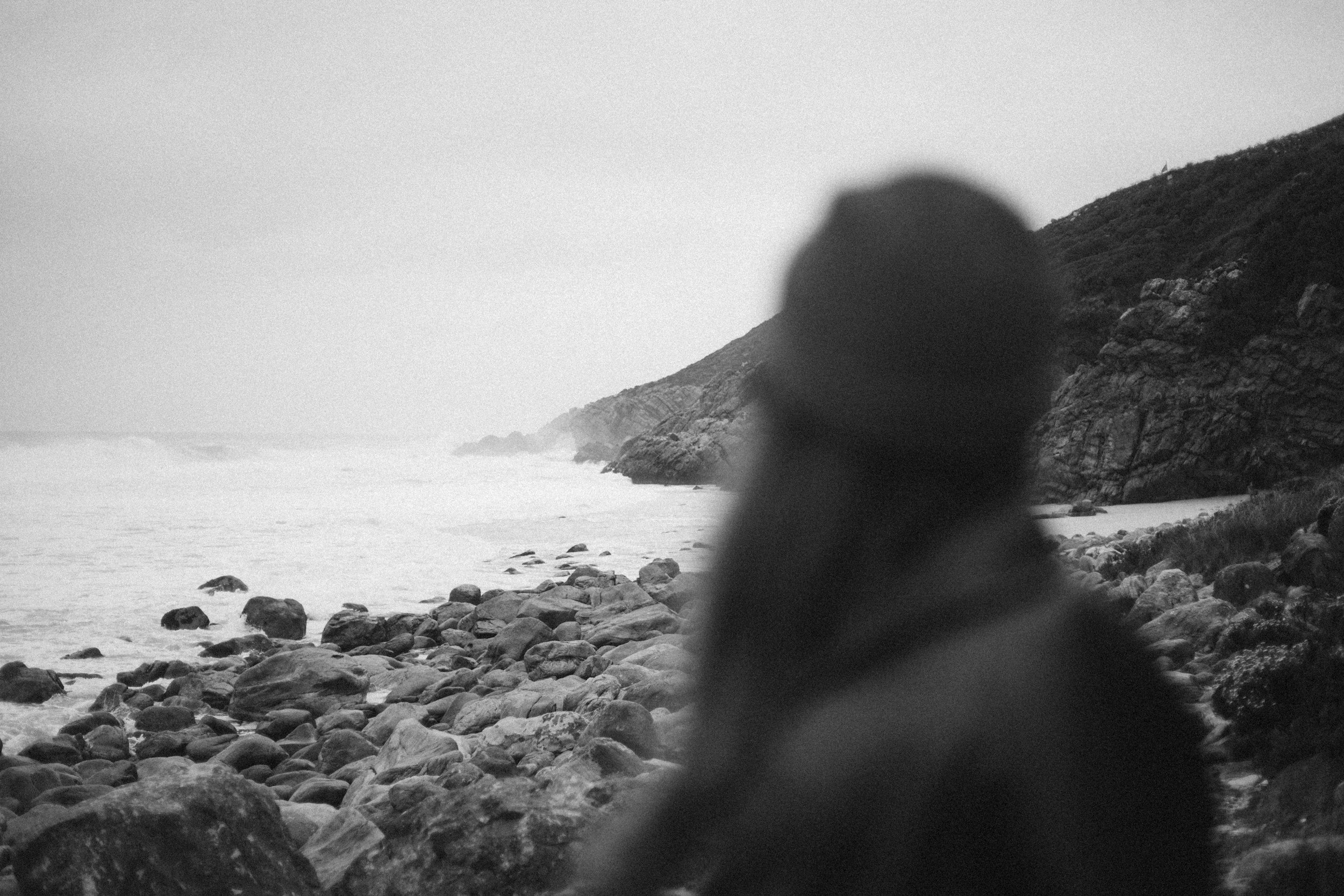 Black and white photo of a rocky coastline with the ocean in the background. A person with dark hair, facing away from the camera, blurred in the foreground.