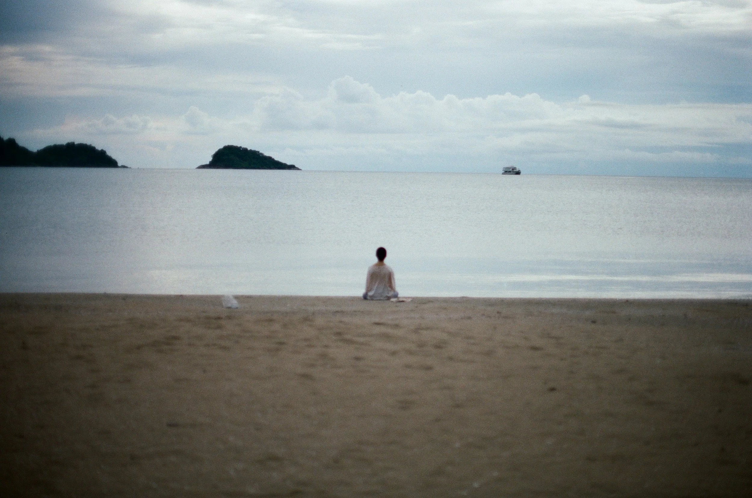 Person sitting on a sandy beach, facing the water, with islands and a boat in the distance under a cloudy sky.