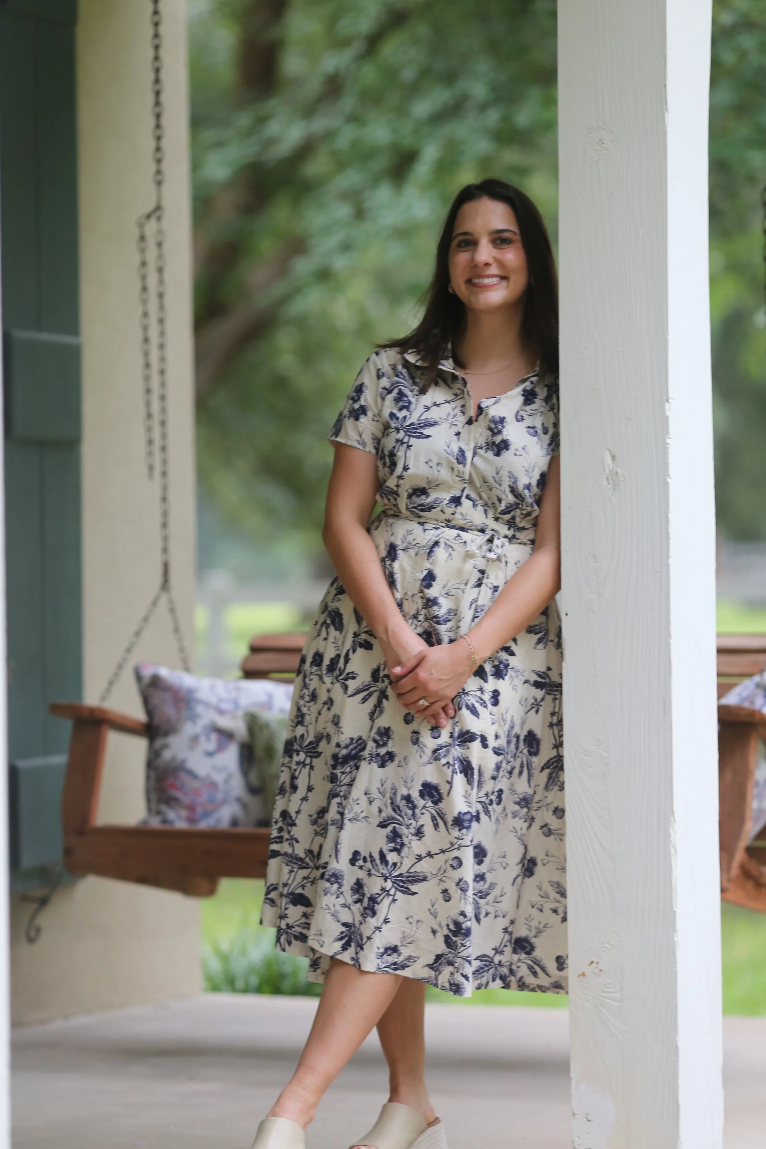 A woman wearing a floral dress standing on a porch, leaning against a white pillar, with a swing and greenery in the background.