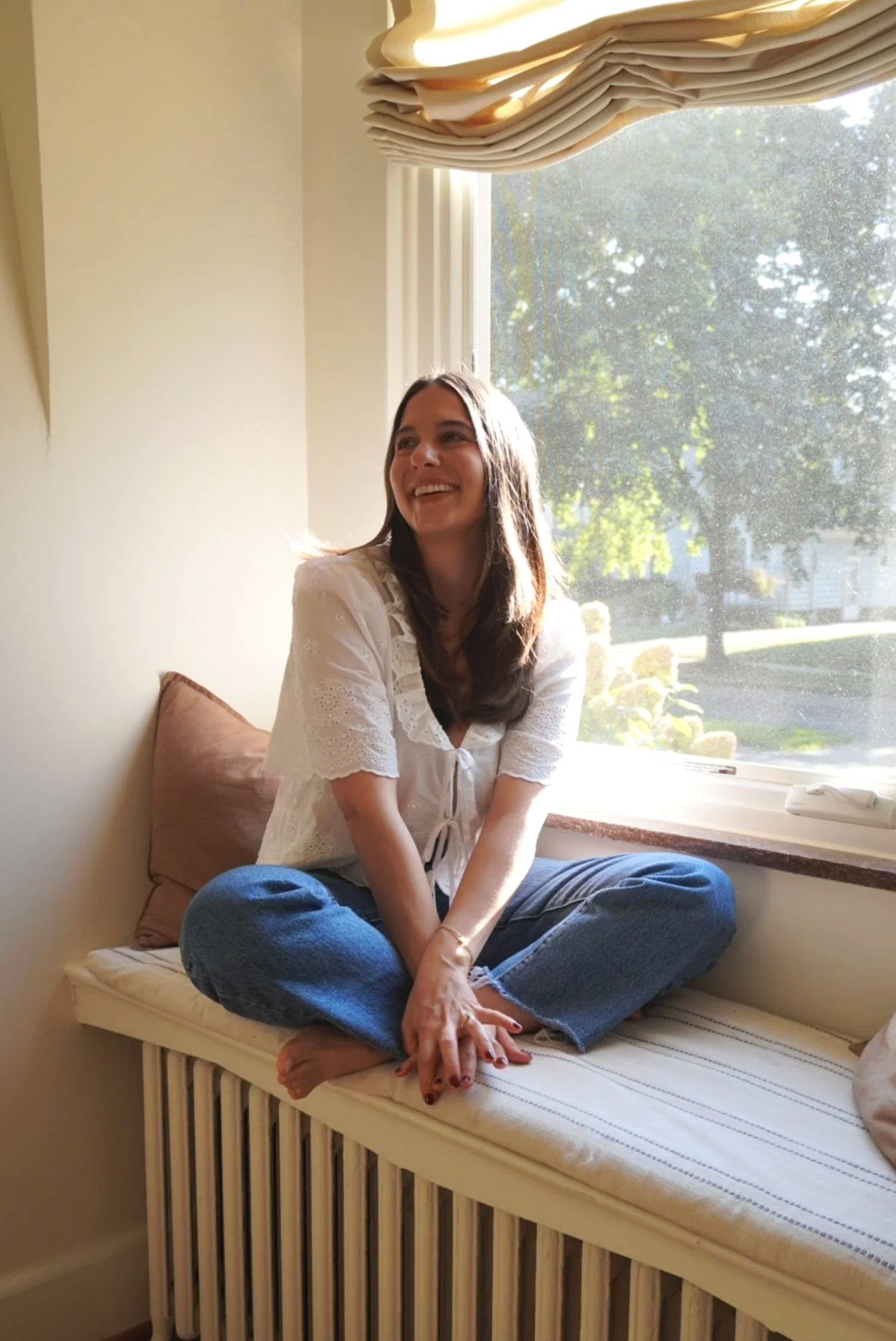 A woman sitting on a window seat, smiling, with sunlight shining in through the window.