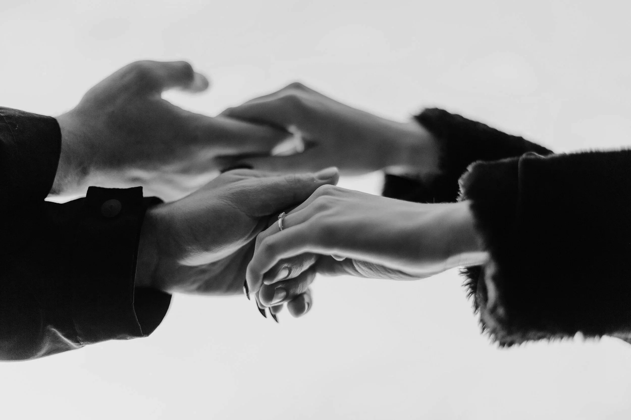 Black and white close-up of two people holding hands with fingers intertwined, one wearing a ring, against a plain light background.