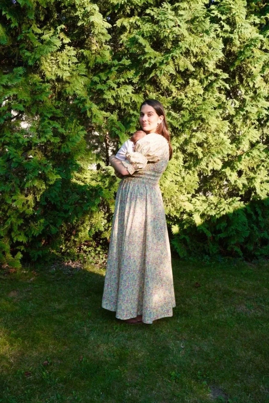 A woman in a long floral dress holding a small baby in her arms, standing on a grassy area with a large green bush behind her.