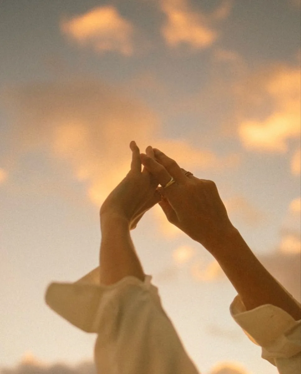 Two hands with rings touching fingers against a sky with soft clouds at sunset.