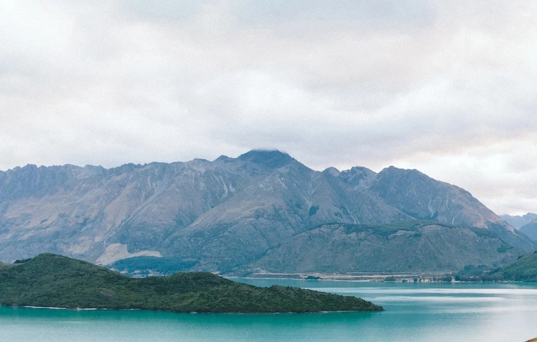 A scenic landscape featuring a large mountain range behind a body of turquoise water and a small, green island in the foreground.