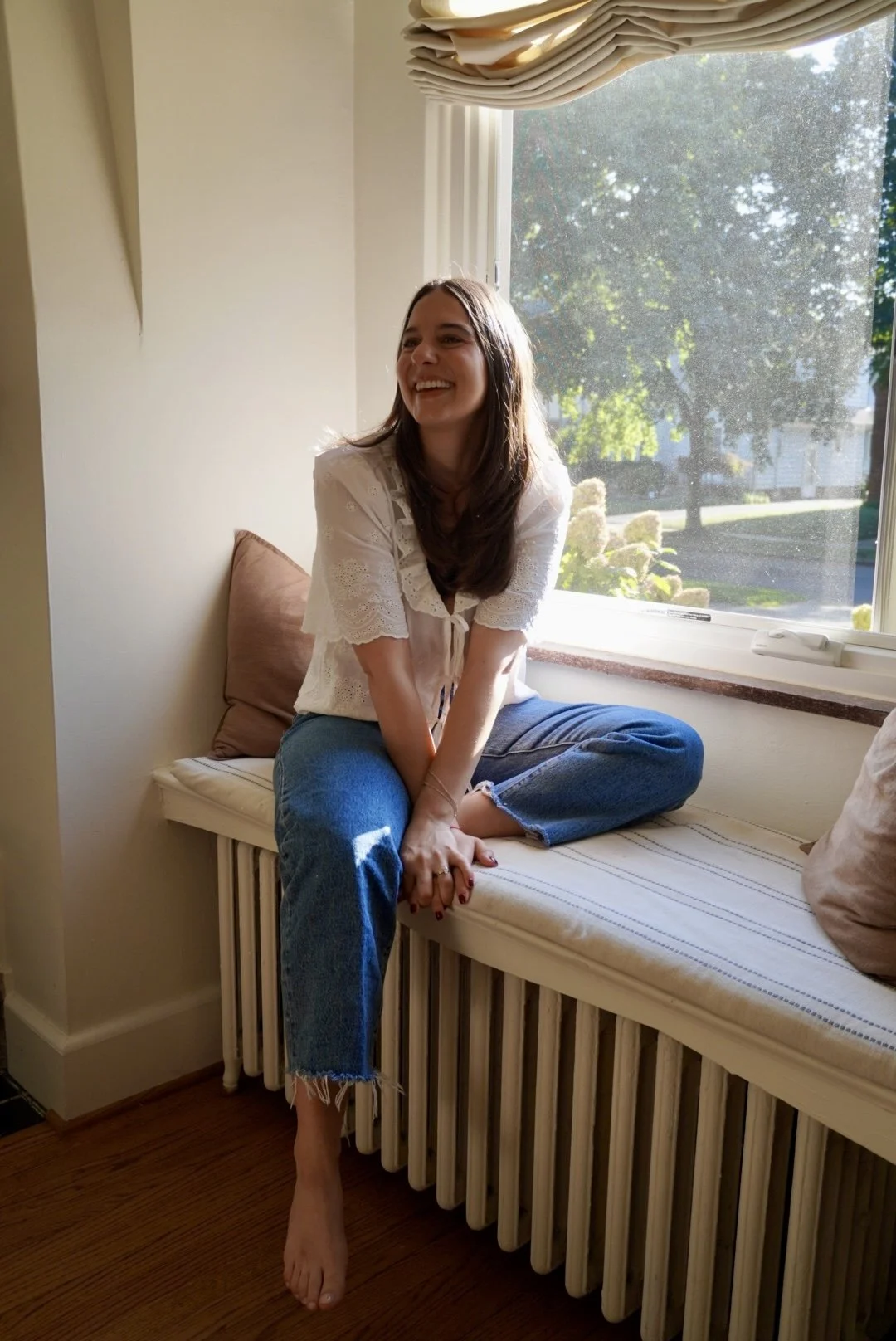 A young woman sitting on a window seat with sunlight streaming in, smiling and looking to the side. She is wearing a white top and blue jeans, with one foot on the floor and the other tucked under her. There are cushions and a view of trees outside the window.