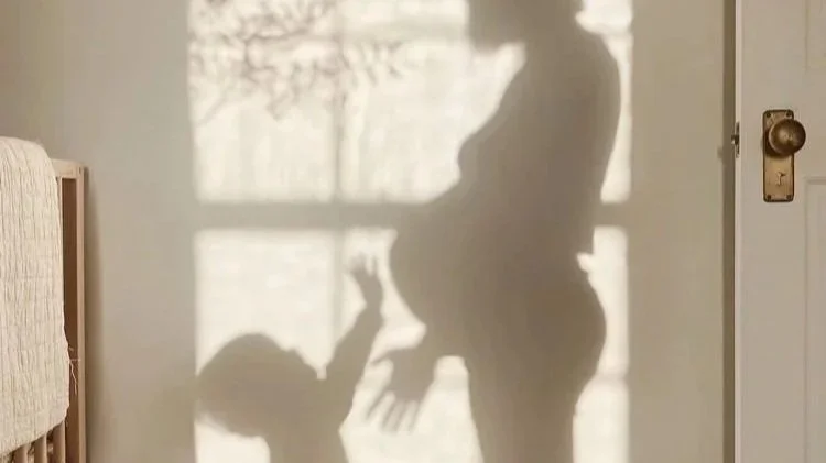 Shadow of a pregnant woman and a baby reaching up towards her is cast on a wall next to a window.
