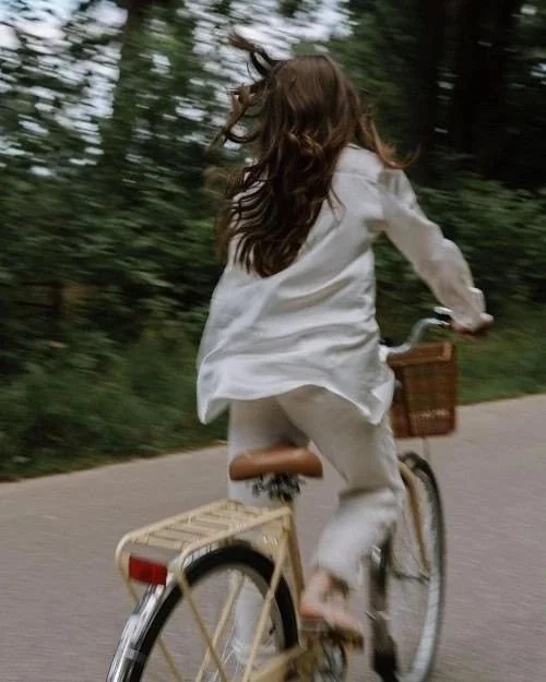 A girl riding a bicycle on a paved path through a wooded area, wearing a white dress with long hair flowing behind her.
